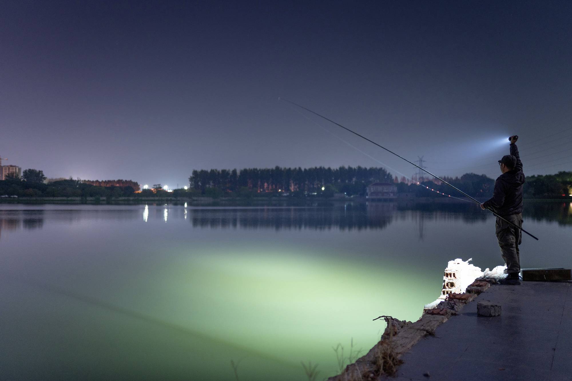 Einzelner Angler steht nachts am See, hält eine Angelrute. Wasser reflektiert Licht, ruhige Atmosphäre.