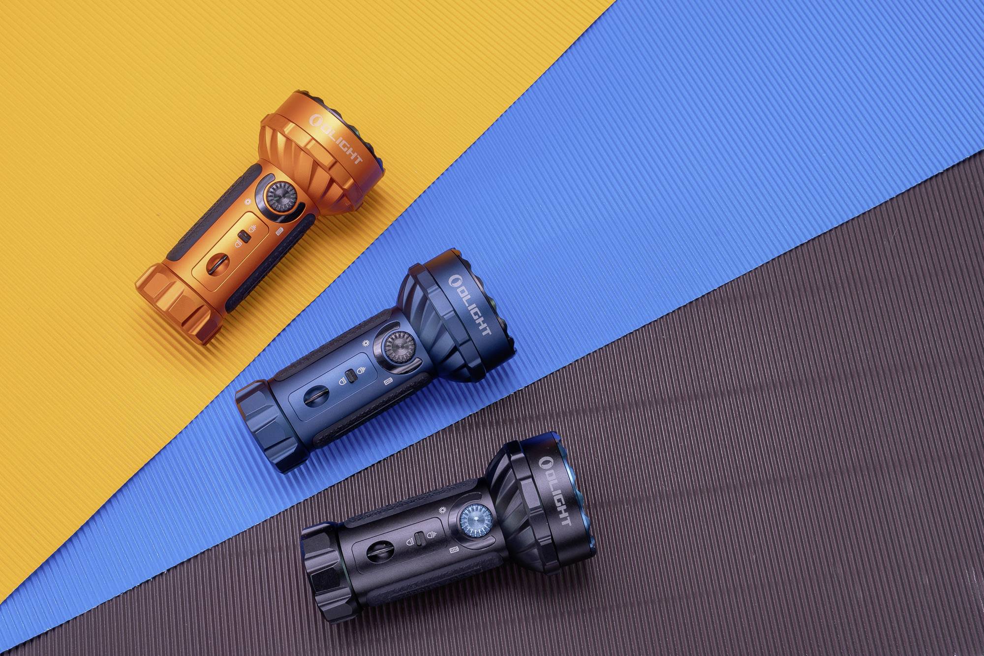 Drei Taschenlampen in Orange, Blau und Schwarz liegen auf bunten Oberflächen in Gelb, Blau und Braun.