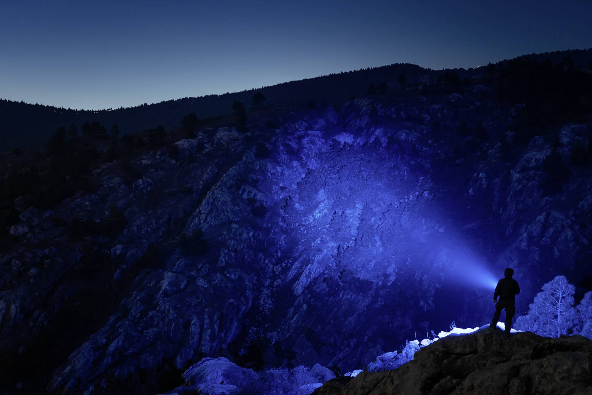 Eine Person steht auf einem Felsen bei Nacht und leuchtet mit einer Taschenlampe in blaues Licht auf eine dunkle Felswand.