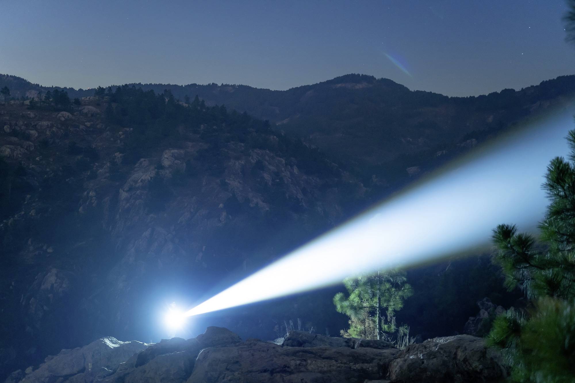Ein starkes Licht strahlt nachts von einem Berg aus in die Ferne, beleuchtet Bäume und Felsen. Dunkler Himmel mit entfernten Hügeln im Hintergrund.