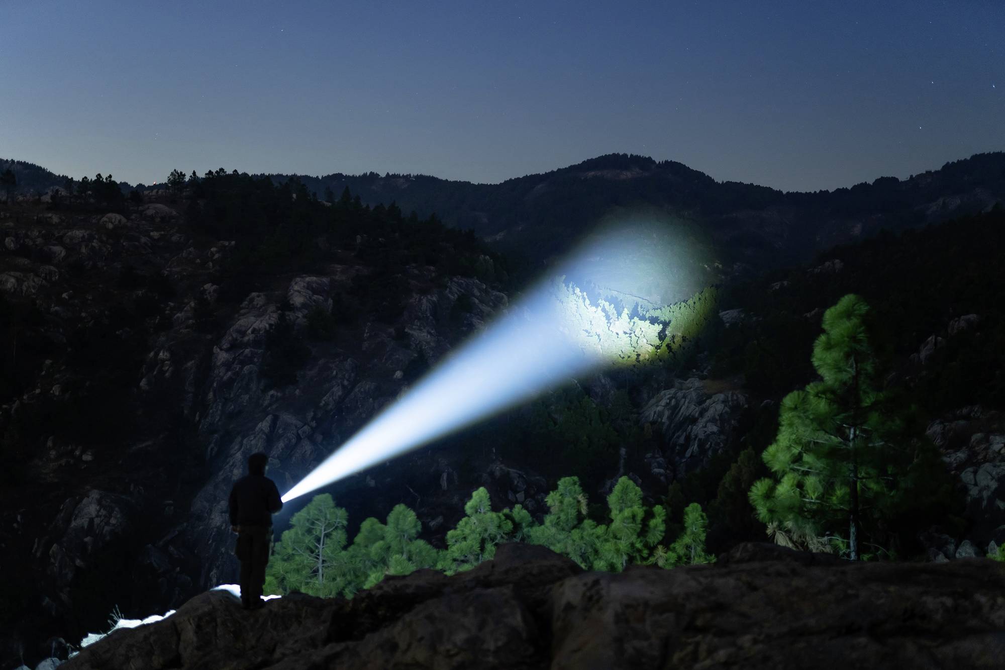 Eine Person leuchtet nachts mit einer starken Taschenlampe auf einen bewaldeten Hügel. Die Umgebung ist von Bergen umgeben.