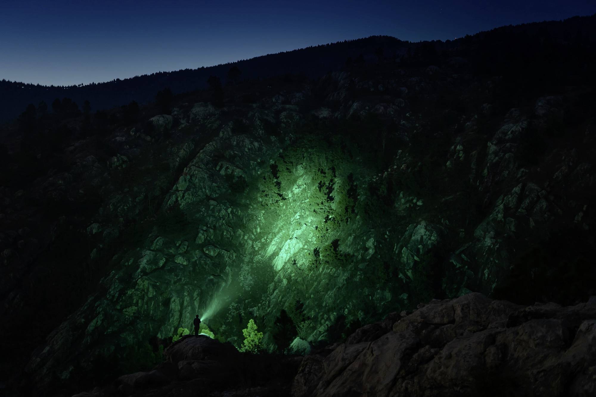 Ein Berglandschaft bei Nacht, beleuchtet von einem grünen Lichtstrahl, der auf Felsen und Bäume strahlt. Eine Person steht im Vordergrund.