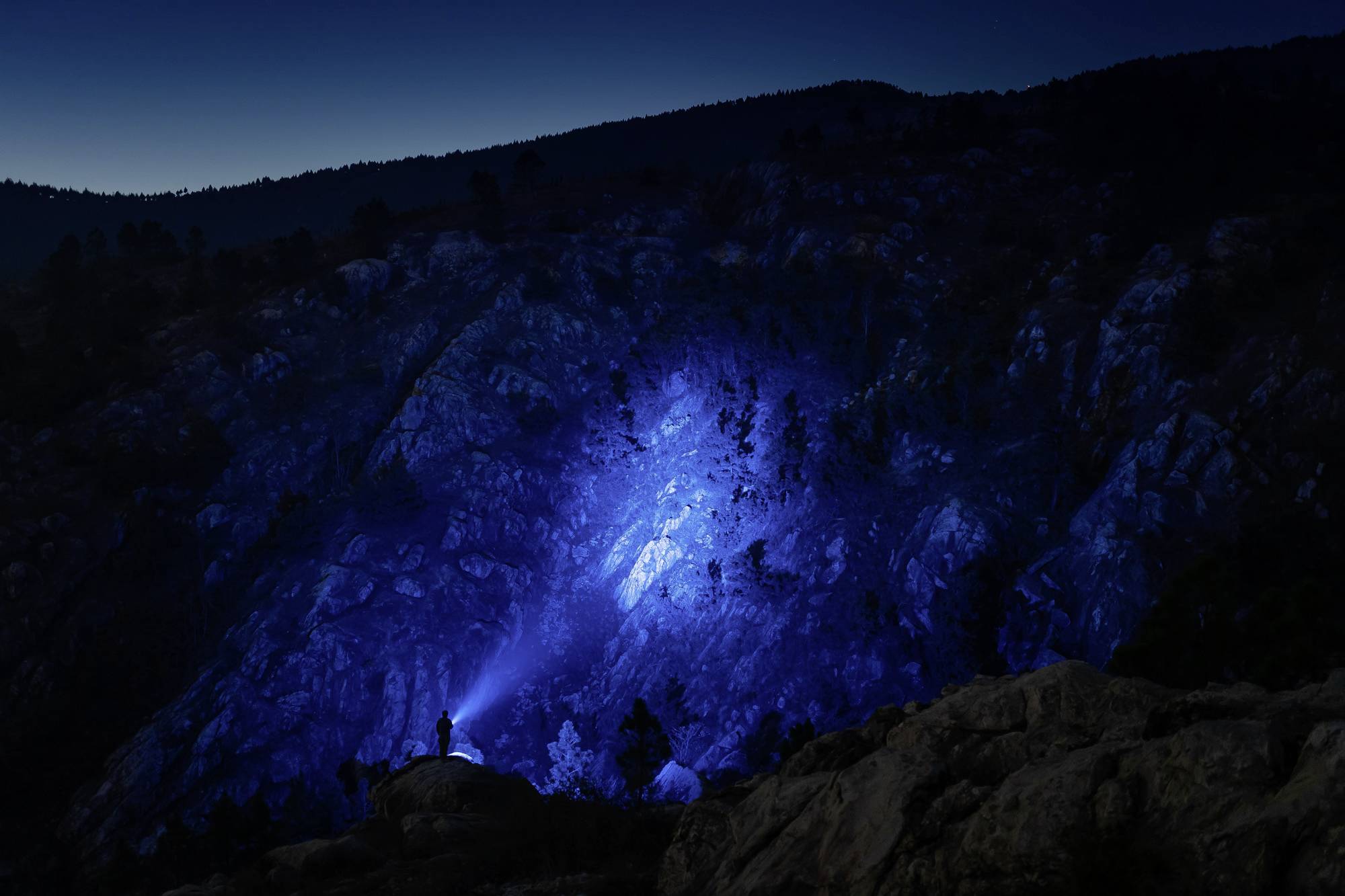 Ein Wanderer beleuchtet mit einer Taschenlampe eine felsige Berglandschaft in der Nacht, wodurch ein blauer Lichtstrahl entsteht.