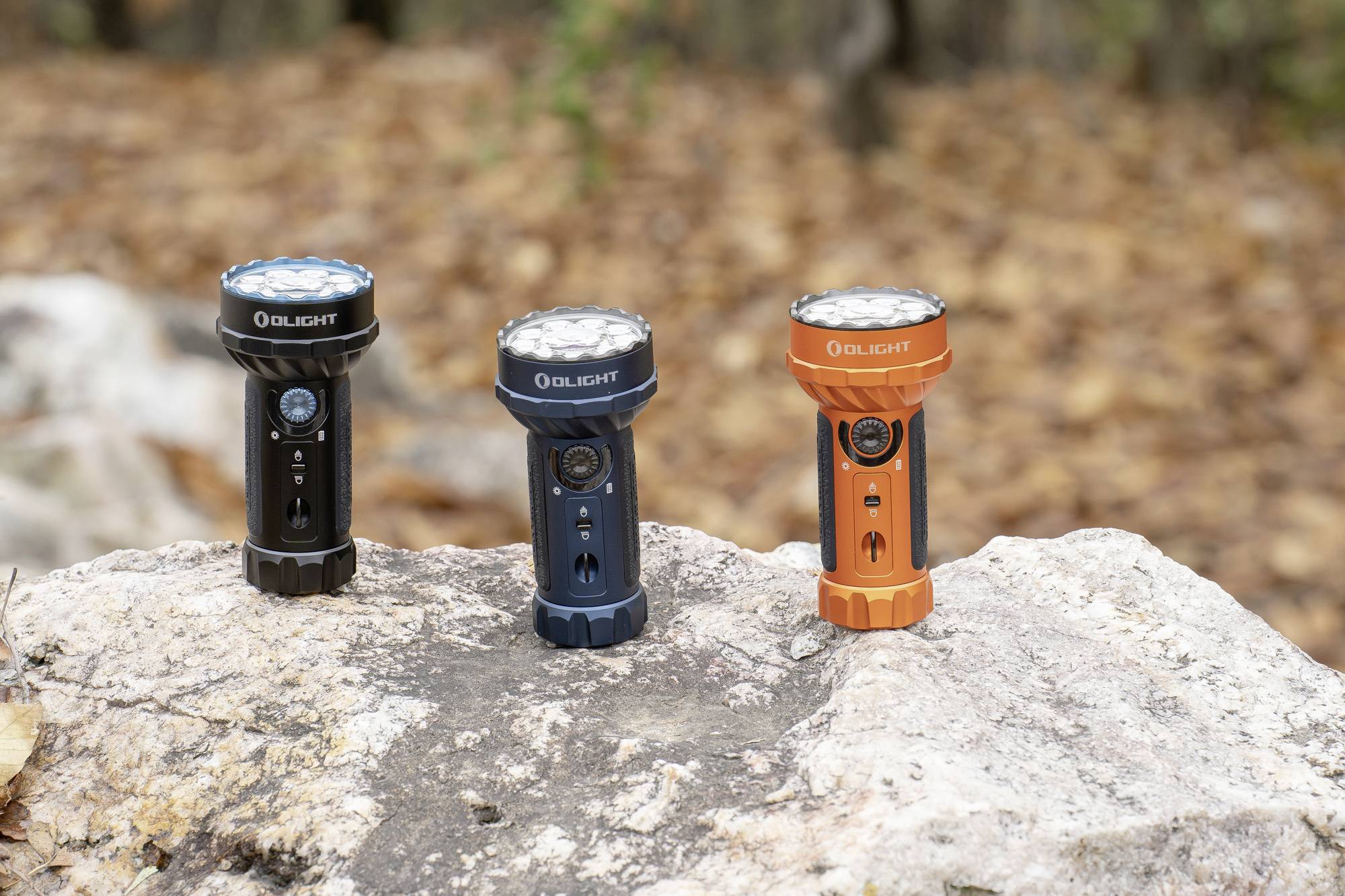 Drei Taschenlampen stehen auf einem Felsen im Freien, umgeben von Herbstlaub. Zwei sind schwarz, eine ist orange. Sie zeigen unterschiedliche Designs.