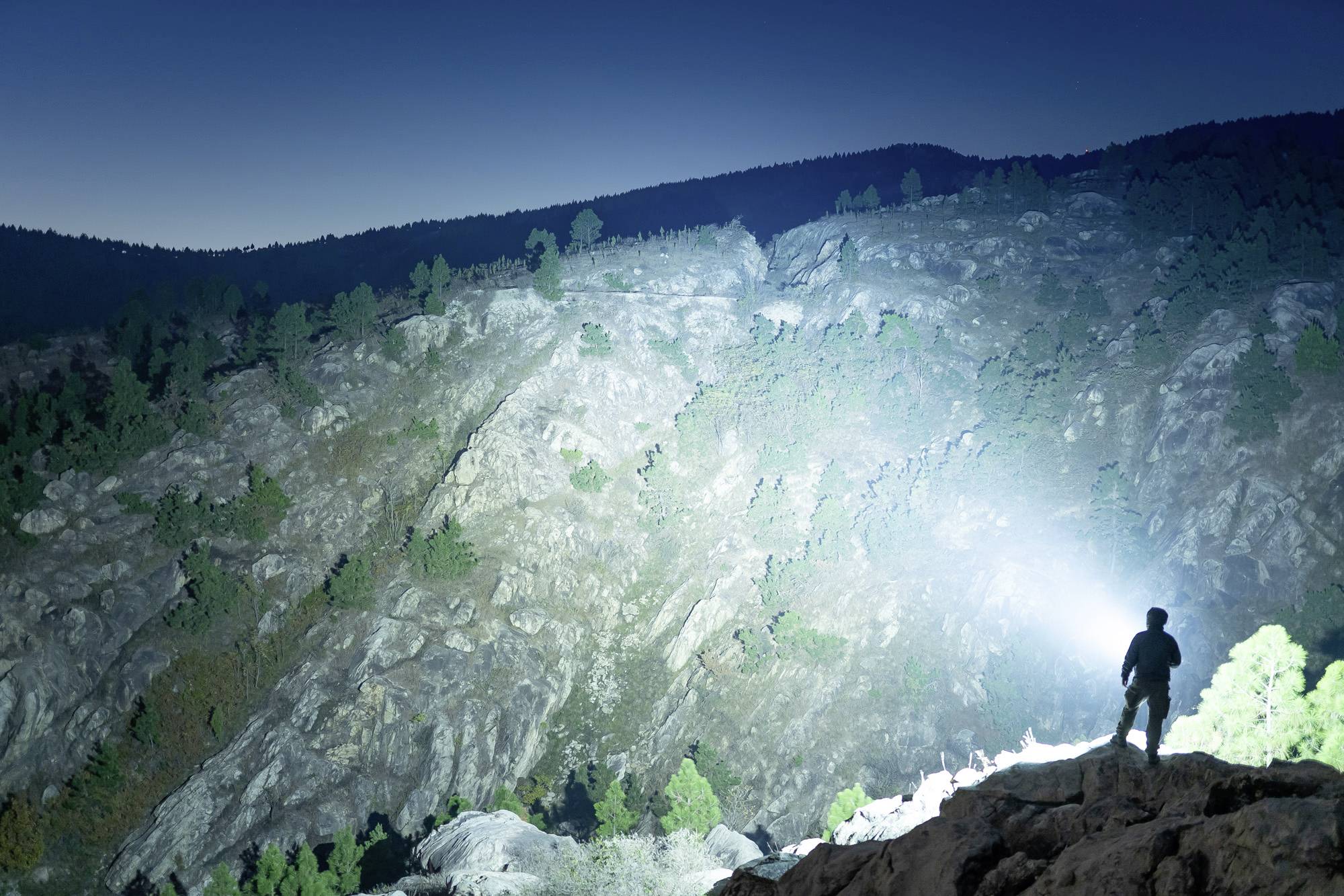 Eine Person steht nachts auf einem beleuchteten Felsen und blickt mit einer Taschenlampe in eine felsige Landschaft.