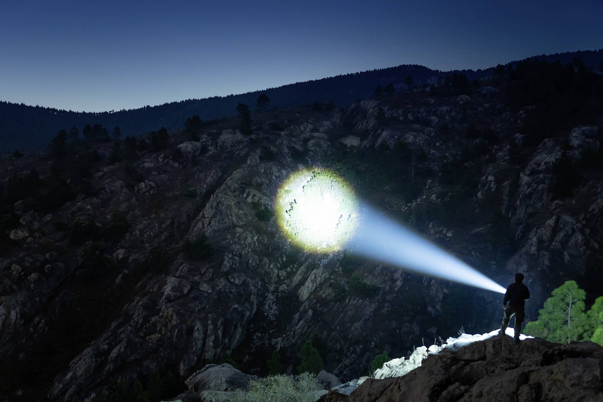 Eine Person beleuchtet bei Nacht eine Berglandschaft mit einer starken Taschenlampe, die einen kreisförmigen Lichtstrahl erzeugt.
