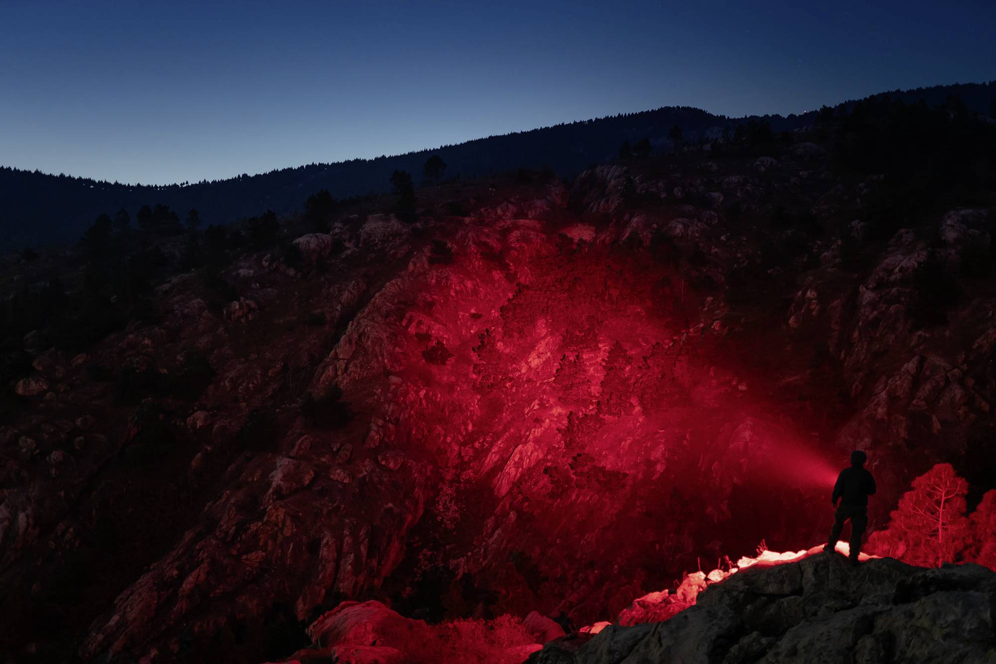 Eine Person steht auf einem Felsvorsprung und leuchtet mit einer roten Taschenlampe auf eine felsige Berglandschaft in der Dämmerung.