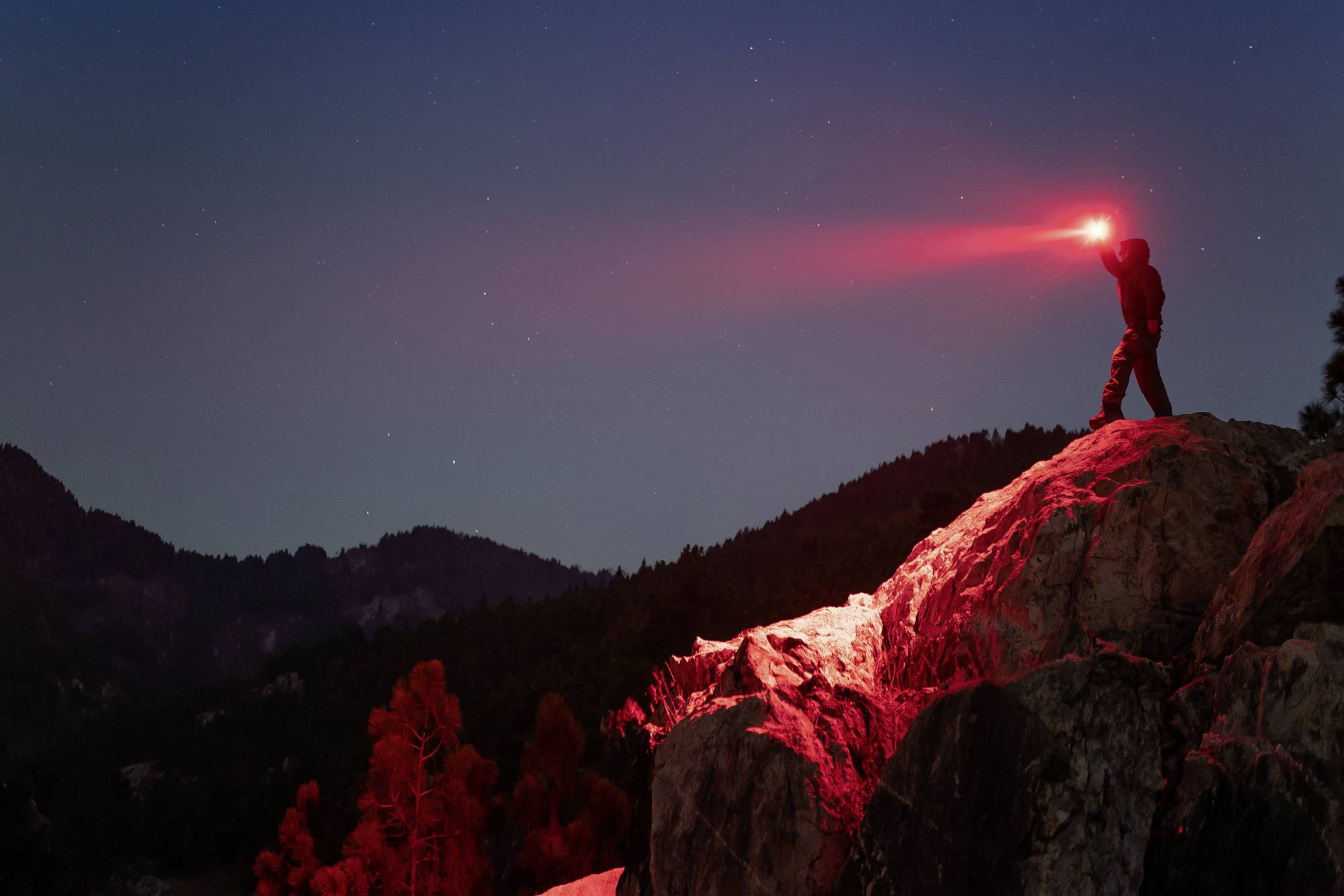 Person auf Felsen bei Nacht, hält eine rote Taschenlampe. Beleuchtet die Umgebung mit rotem Licht, umgeben von Hügeln und Bäumen.