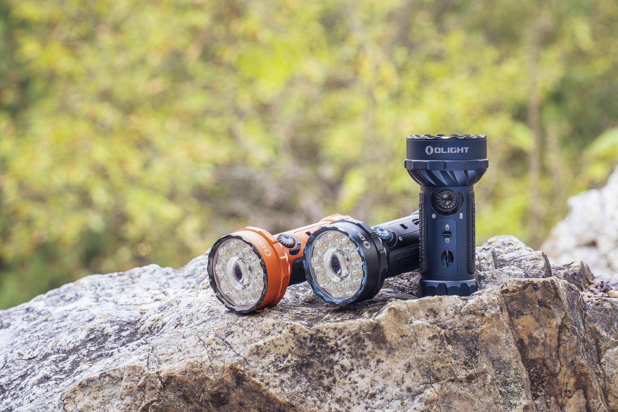 Zwei Taschenlampen stehen nebeneinander auf einem Felsen. Eine ist orange, die andere schwarz. Hintergrund zeigt unscharfen Wald.