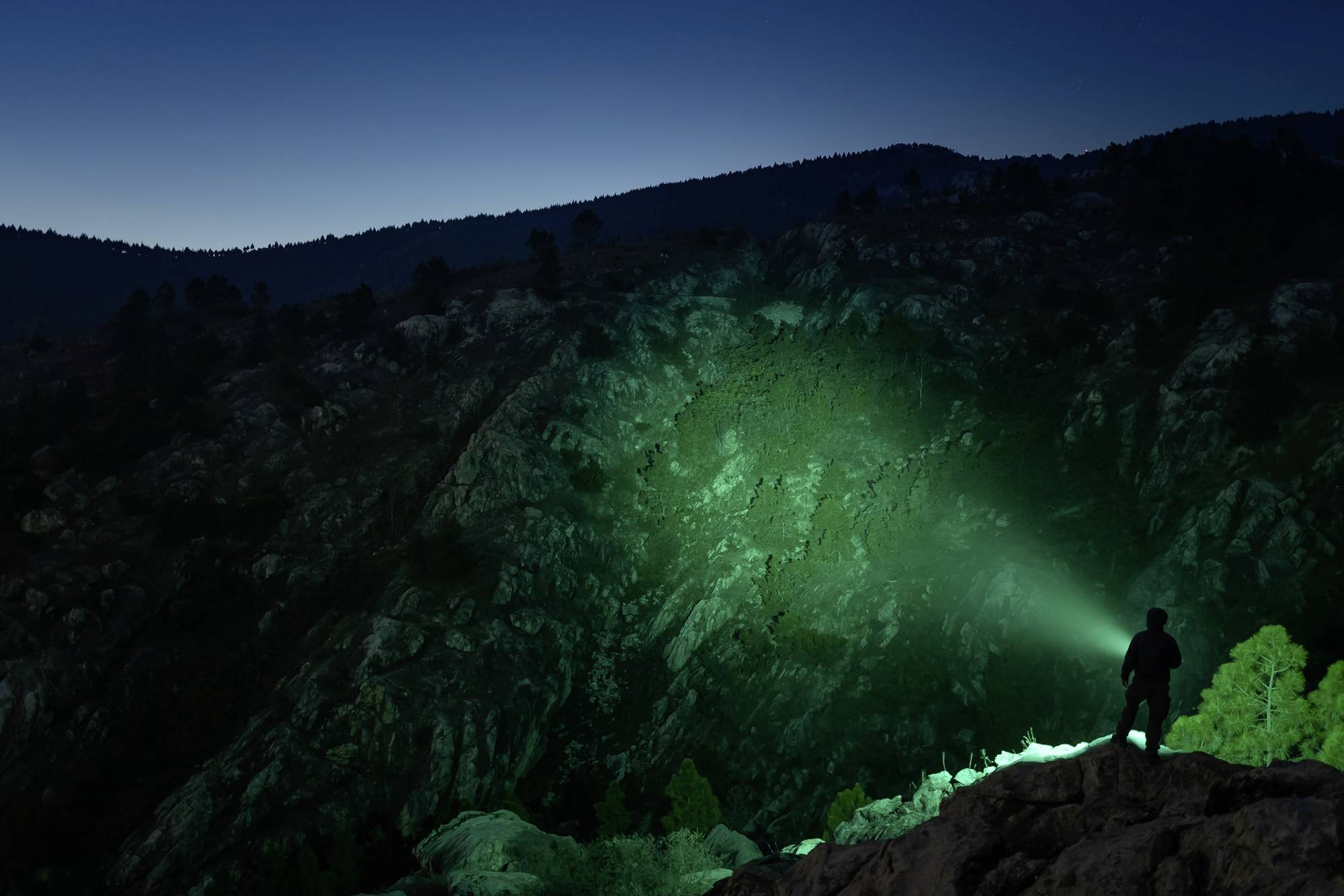 Eine Person steht auf einem Felsen in einer nächtlichen Berglandschaft und leuchtet mit einer Taschenlampe an eine steile Felswand.