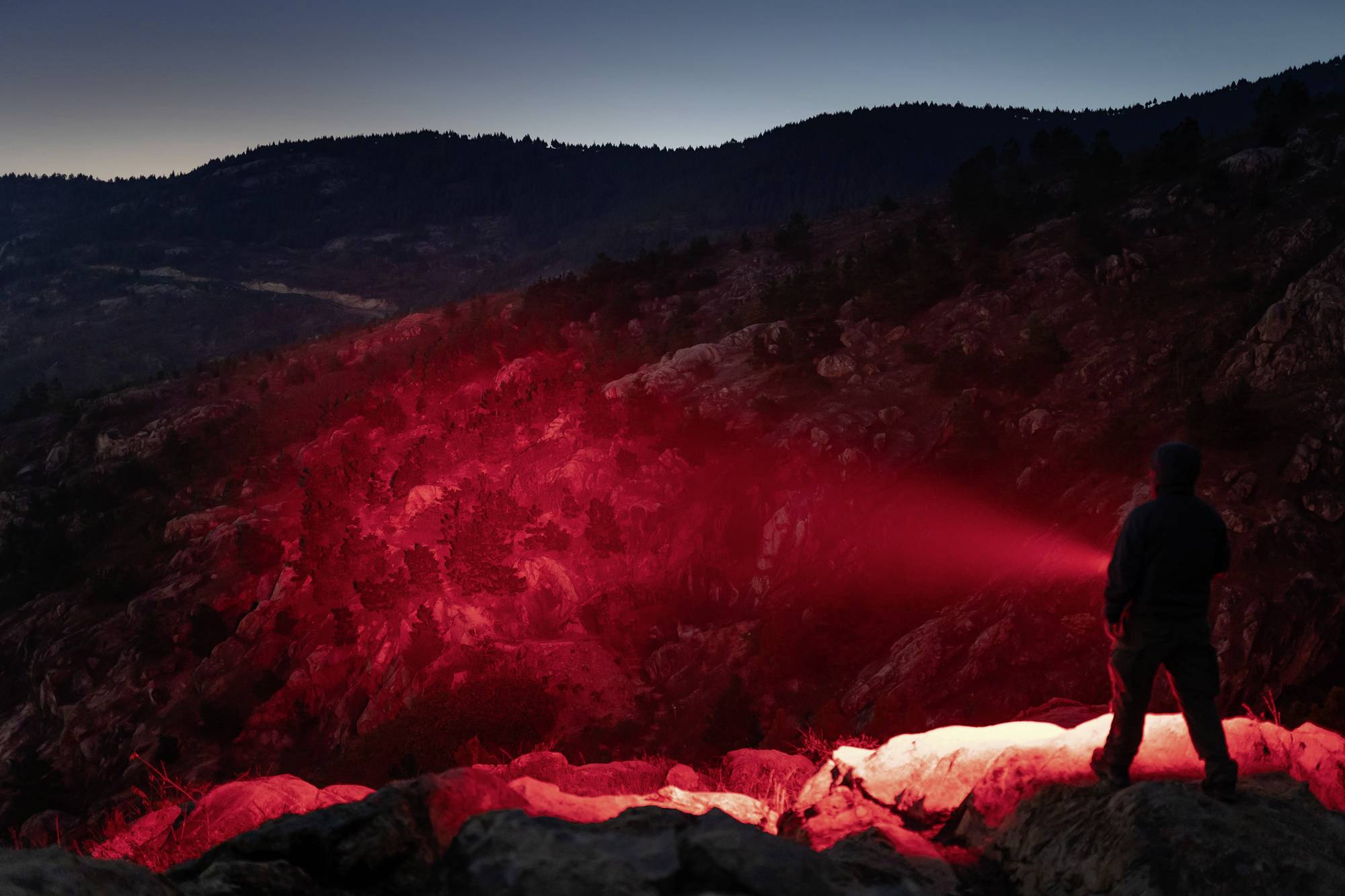 Eine Person steht auf einem Felsen bei Dämmerung und leuchtet mit einer roten Taschenlampe in eine Berglandschaft.