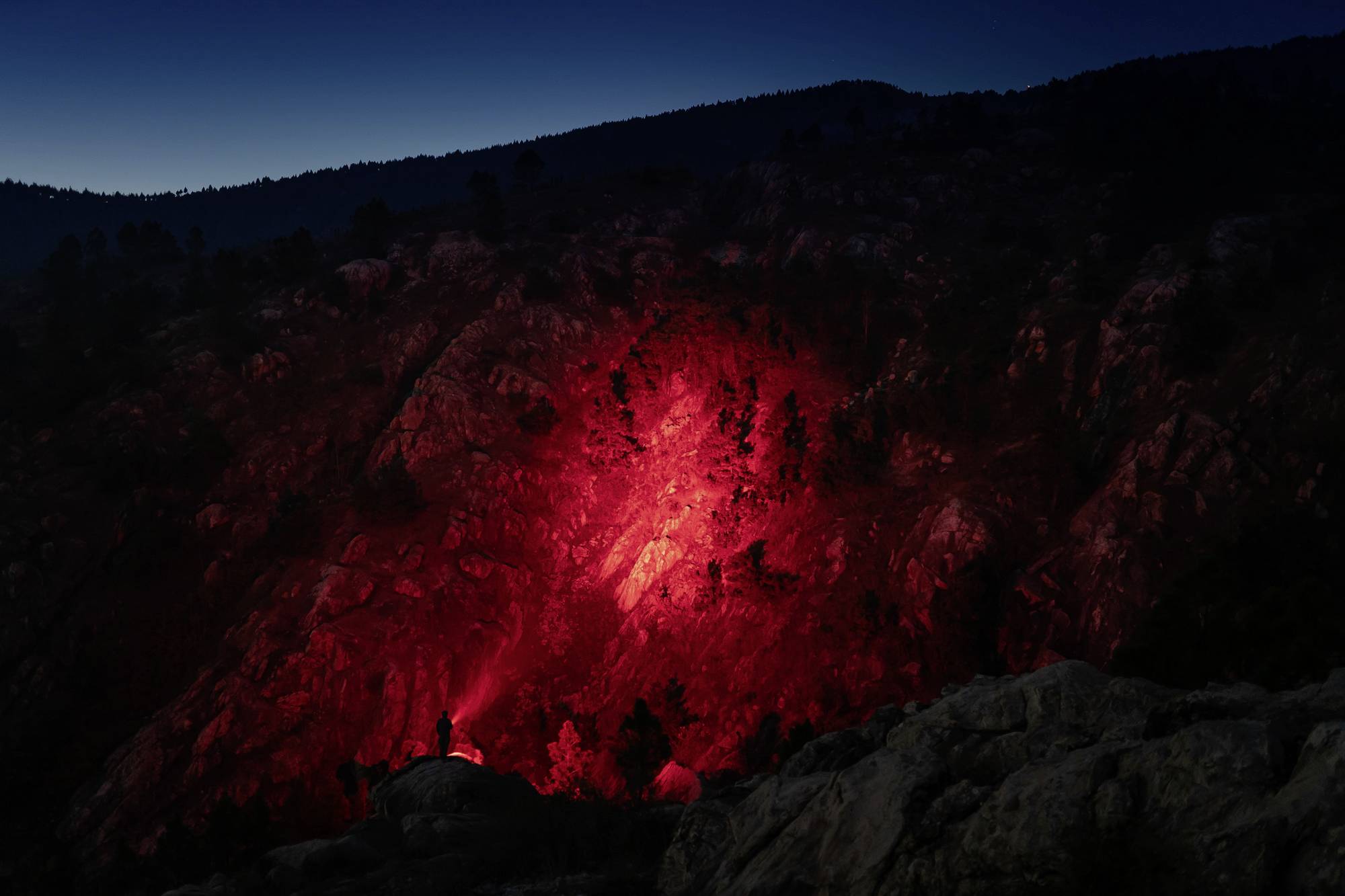 Eine Person steht nachts auf einem Felsen und leuchtet mit einer roten Taschenlampe die umliegenden Berge an.