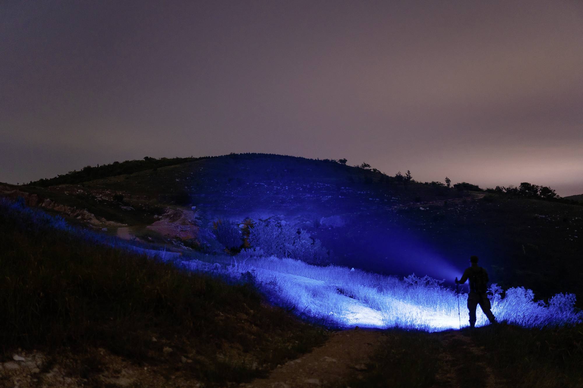 Eine Person steht nachts auf einem Hügel und leuchtet mit einer Taschenlampe, wodurch blaues Licht auf den Boden scheint.