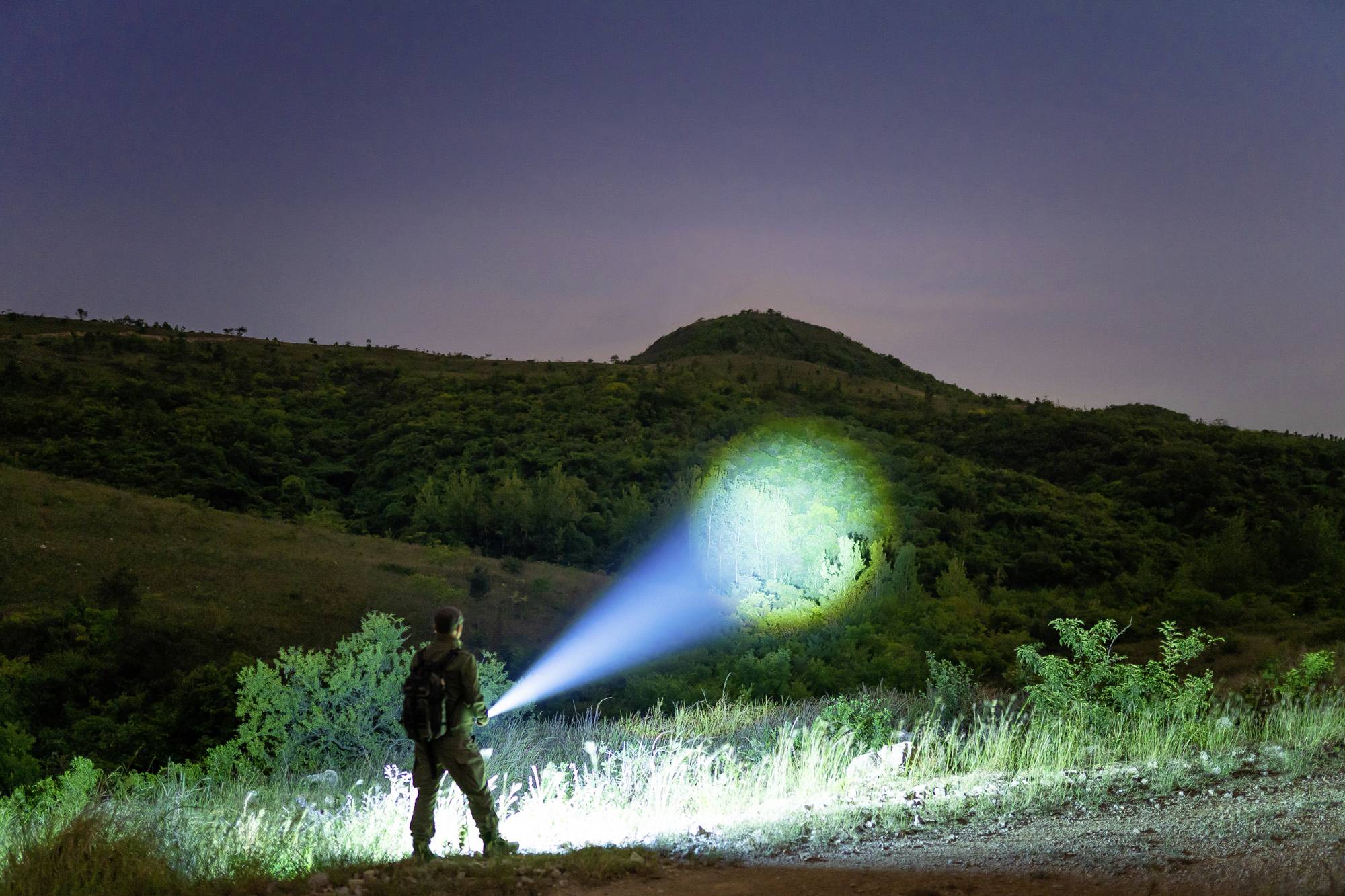 Ein Mensch steht nachts im Freien und leuchtet mit einer Taschenlampe einen Hügel an, der von Büschen und Bäumen umringt ist.