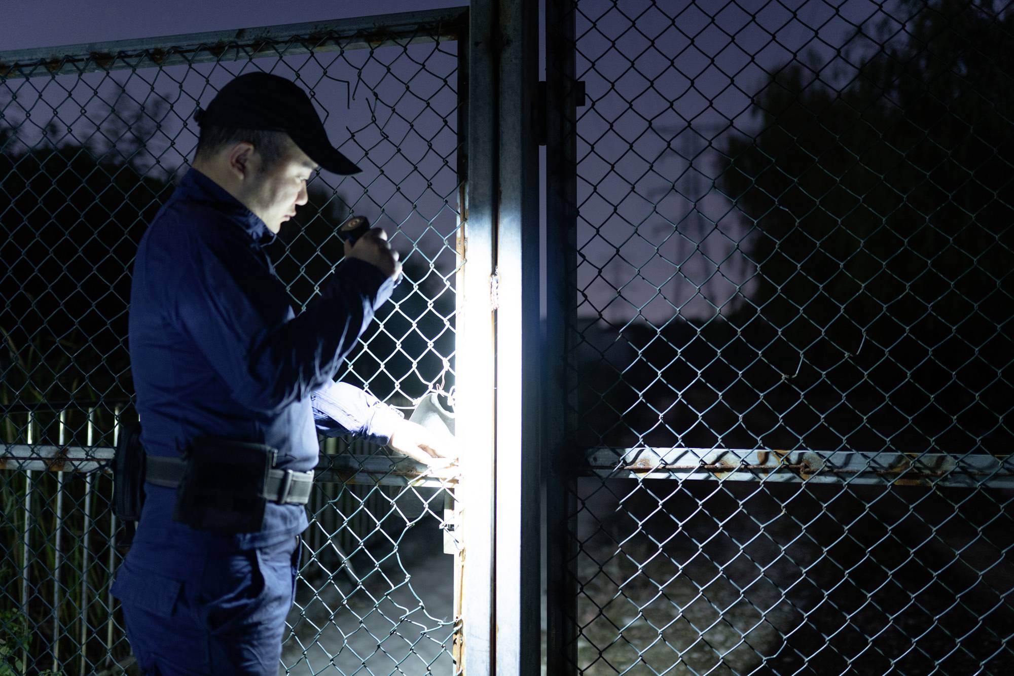 Ein Sicherheitsbeamter untersucht bei Nacht mit einer Taschenlampe ein verschlossenes Metallgittertor.