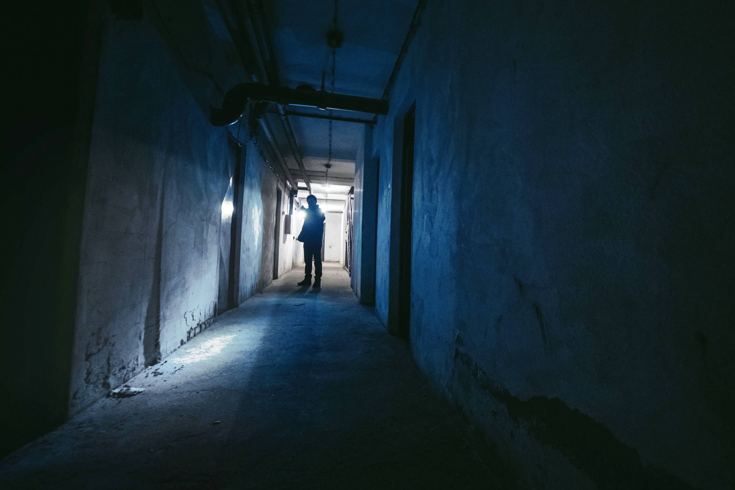 Dunkler, schmaler Korridor mit einer silhouettenhaften Person am Ende, die in Richtung einer schwachen Lichtquelle schaut. Mysteriöses Ambiente.