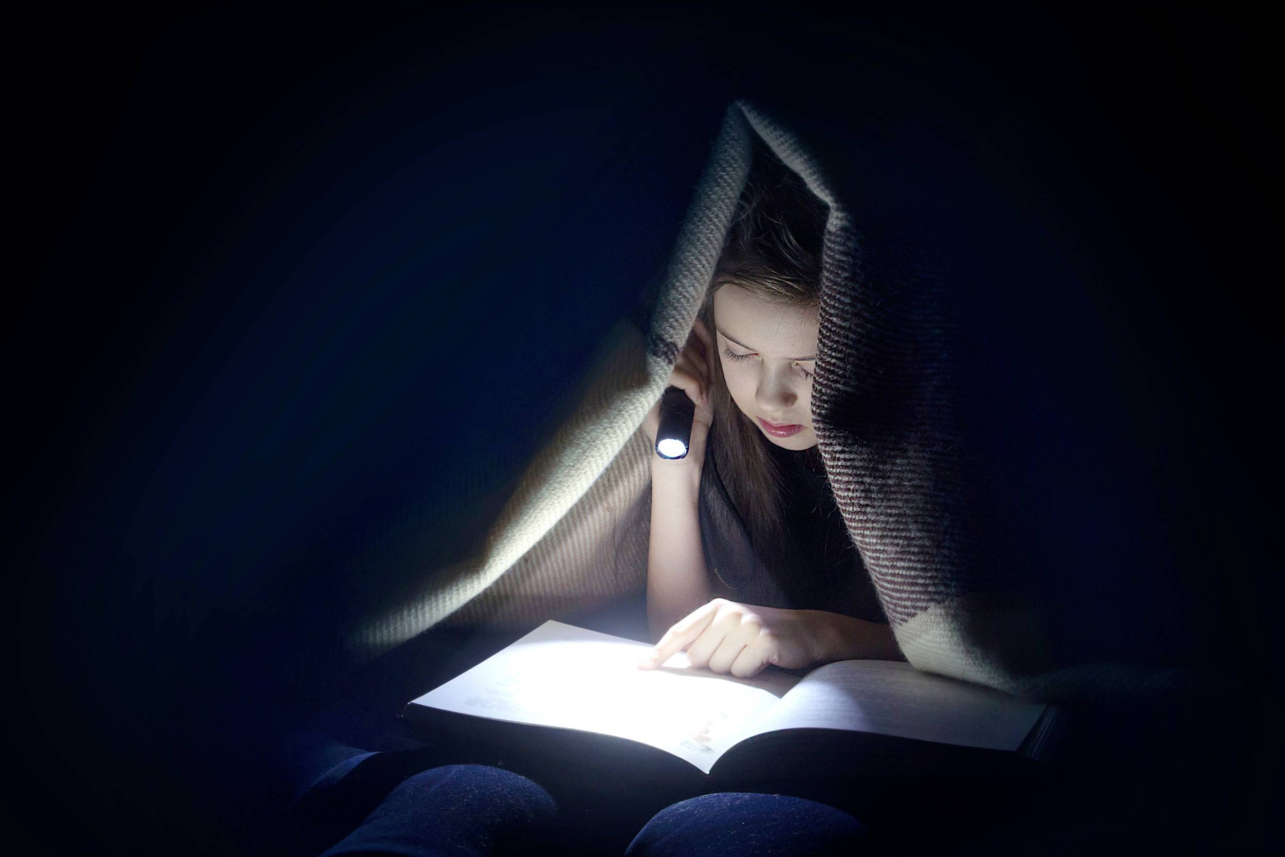 Ein Kind liest heimlich unter einer Decke mit einer Taschenlampe. Es schafft eine geheimnisvolle und konzentrierte Atmosphäre.