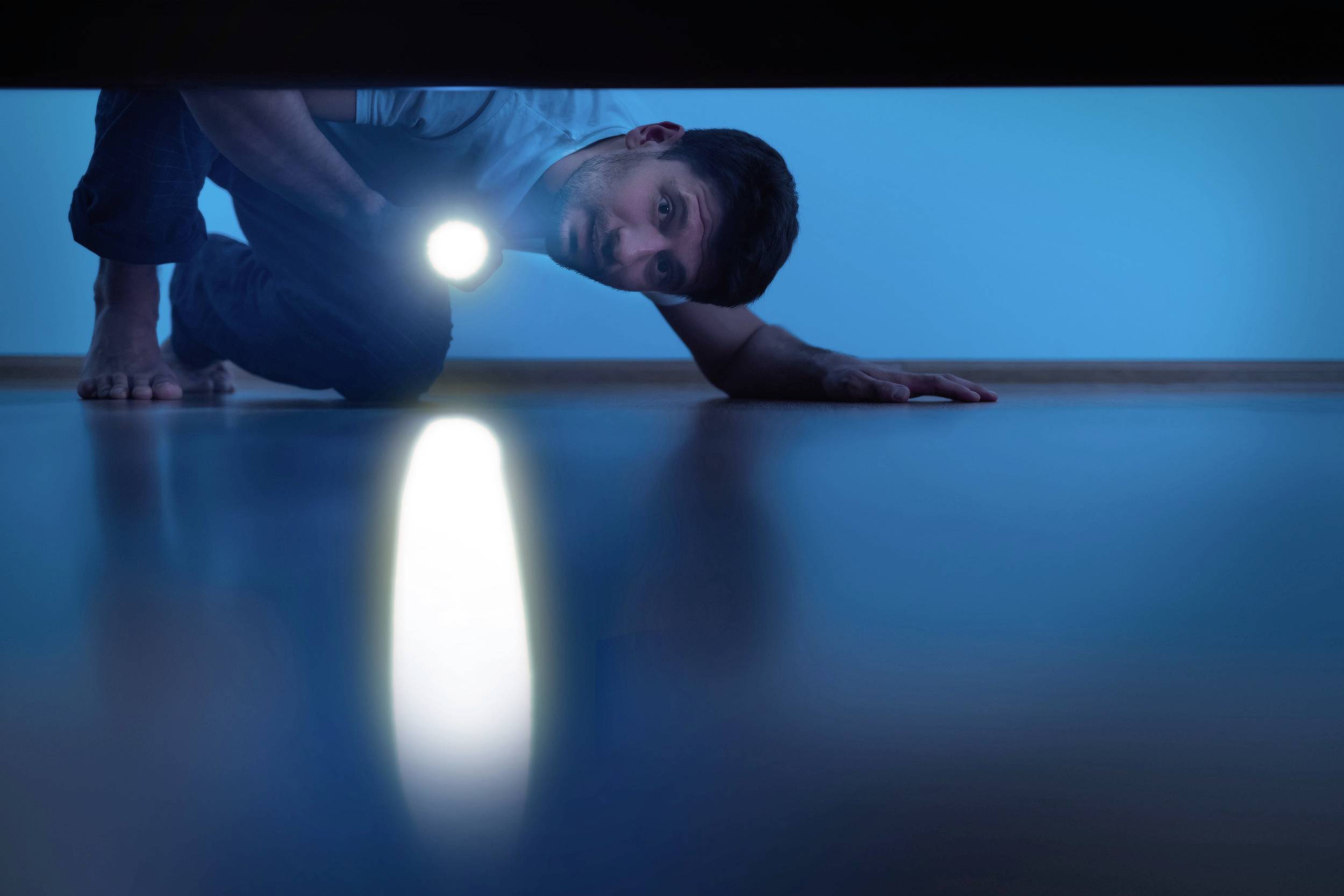 Ein Mann leuchtet mit einer Taschenlampe unter ein Bett. Sein Gesicht und seine Hand sind sichtbar. Der Raum ist blau beleuchtet.