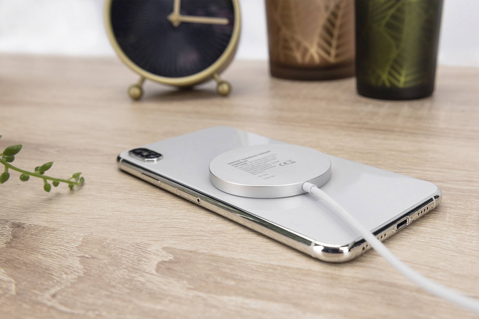 Ein drahtloses Ladegerät liegt auf einem Smartphone, das auf einem Holztisch neben einer Uhr, einer Pflanze und Dekorationsgegenständen liegt.