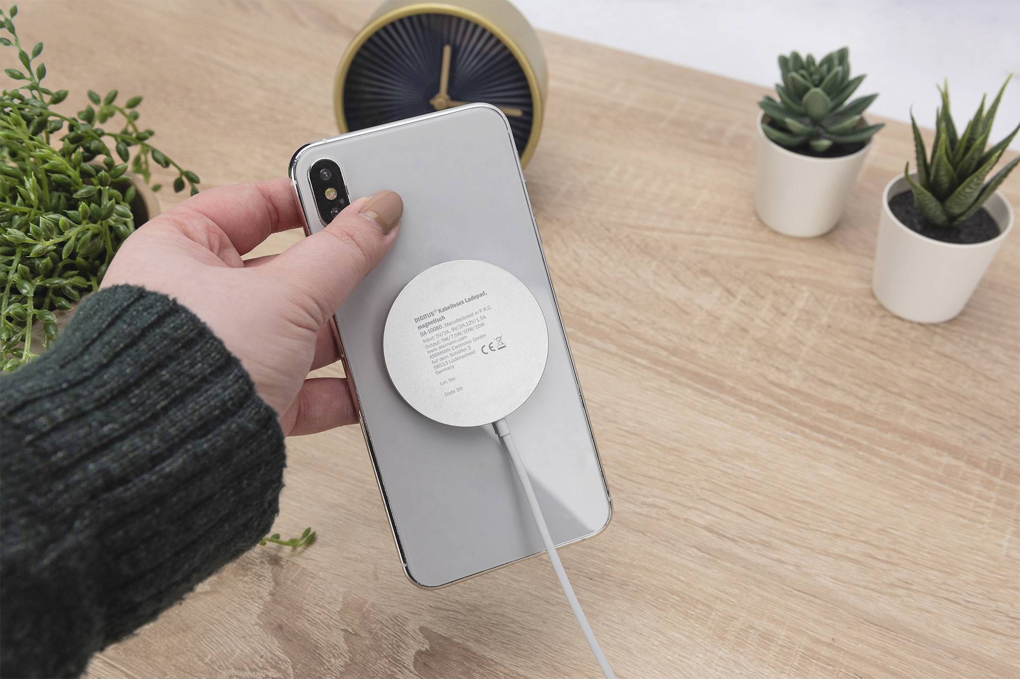 Eine Person hält ein Smartphone mit einer runden drahtlosen Ladepads, das an einem Gerät befestigt ist. Die Umgebung umfasst kleine Topfpflanzen auf einer Holzoberfläche.