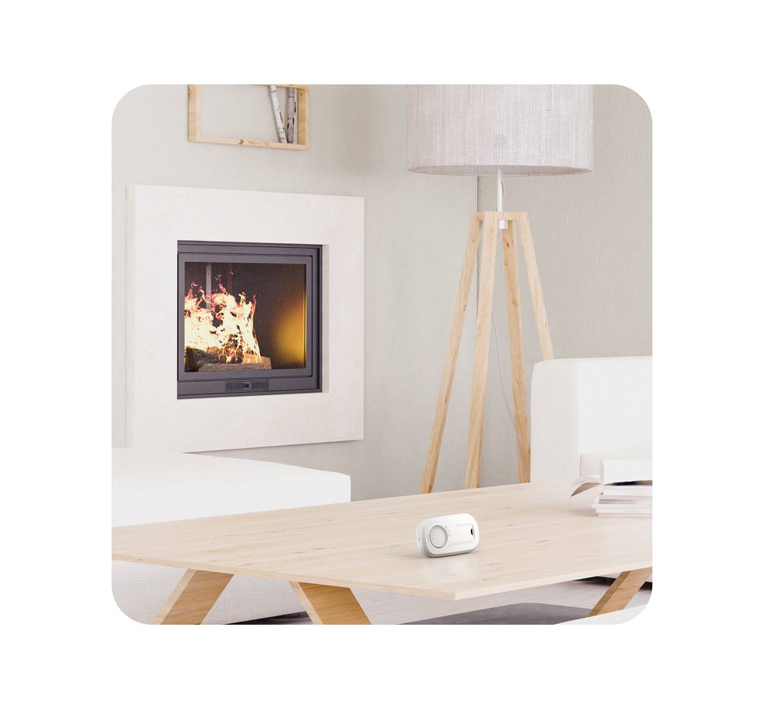 Ein modernes Wohnzimmer mit einem brennenden Kamin, einem weißen Sofa, einem Holztisch und einer Stehlampe. Auf dem Tisch liegt ein kleines weißes Gerät.