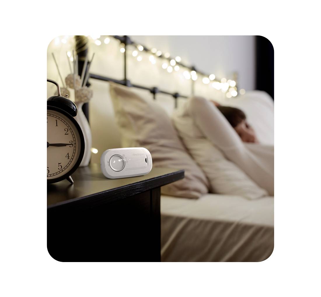 Eine Person liegt entspannt auf einem Bett im Hintergrund, während im Vordergrund ein Wecker und ein elektronisches Gerät auf einem Tisch stehen.