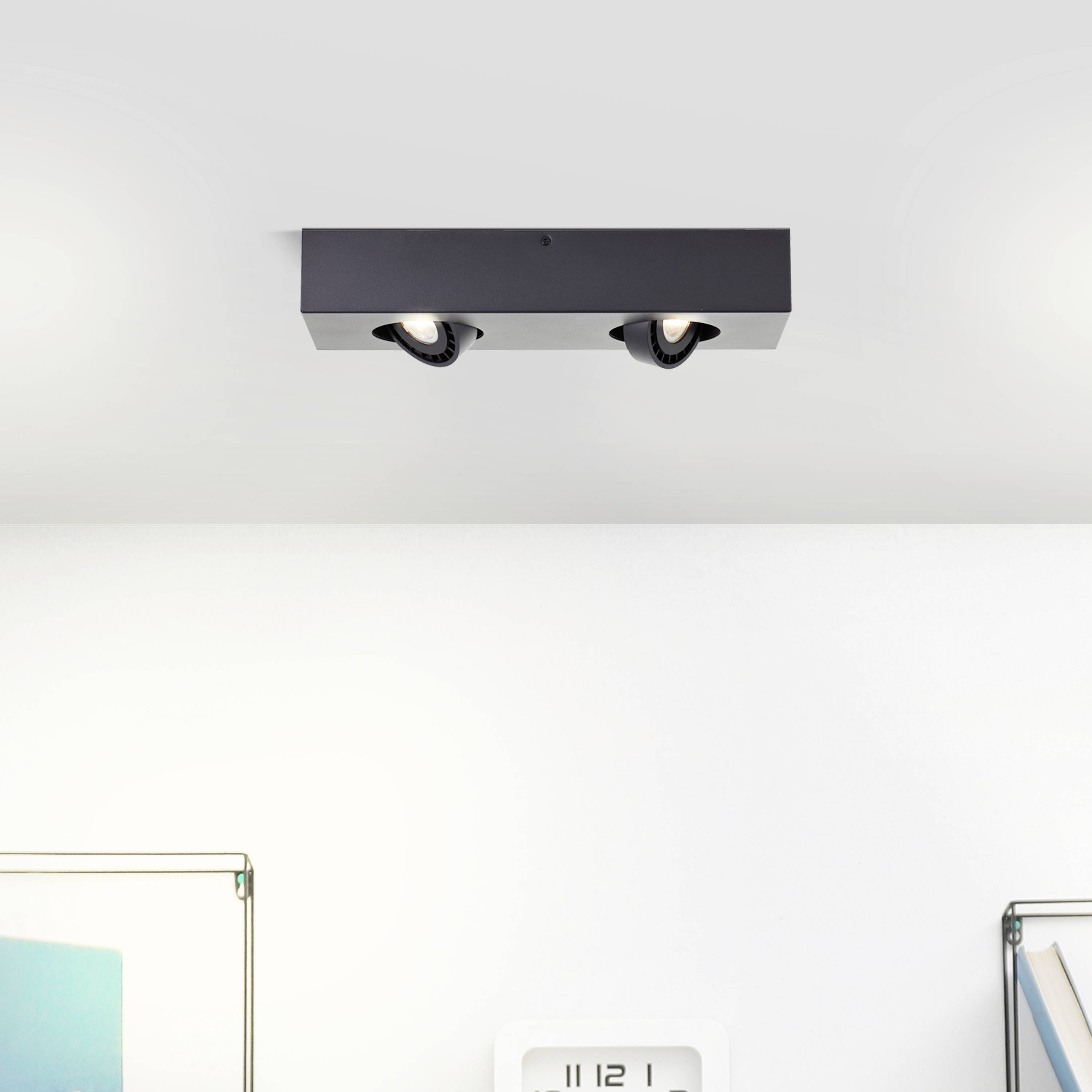 Ein schwarzes Deckenlicht mit zwei runden Lampen ist an einer weißen Decke montiert. Darunter ist die Ecke eines Raumes sichtbar.