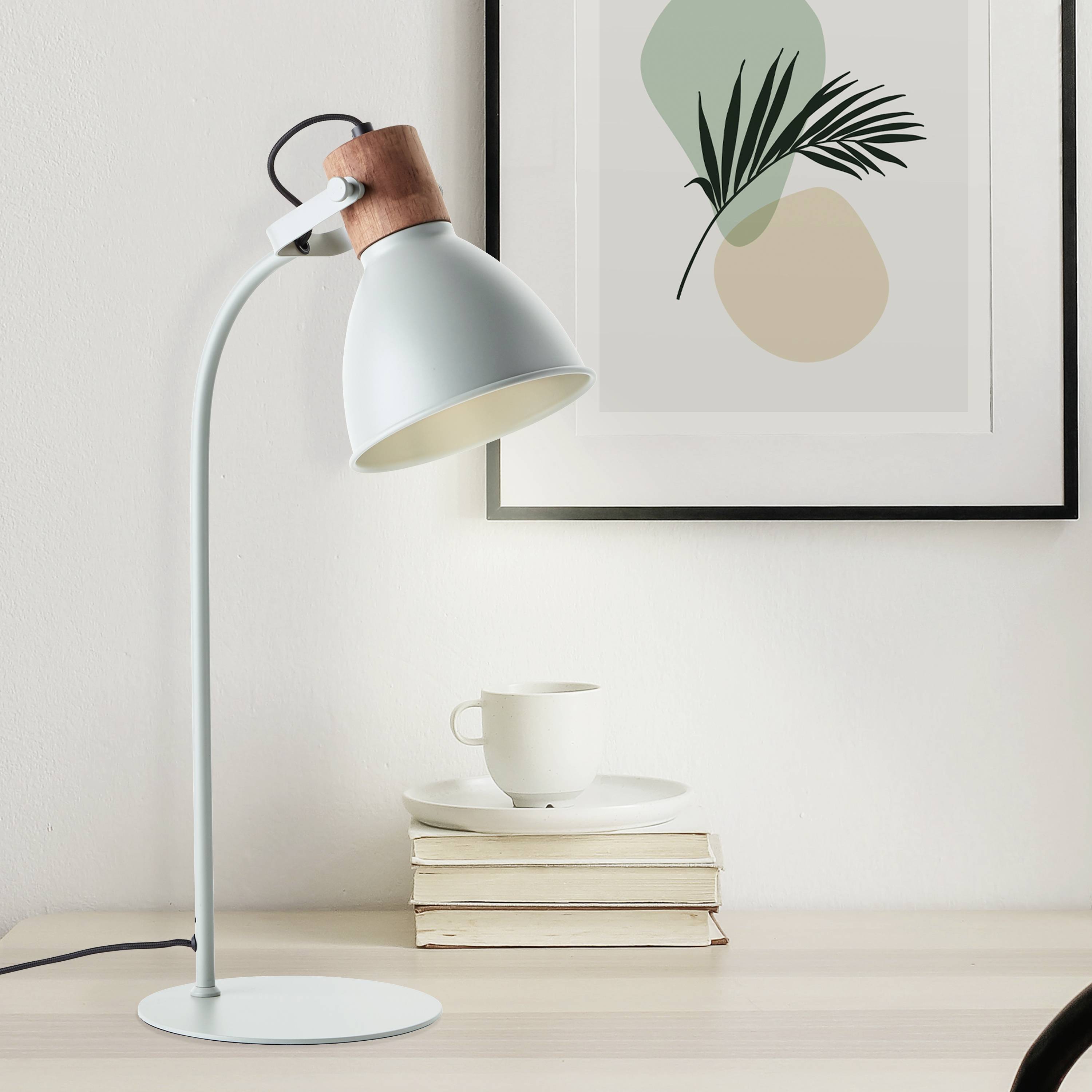 Ein moderner Schreibtisch mit einer weißen Lampe links, gestapelten Büchern, einer Tasse und einem minimalistischen Kunstbild an der Wand.