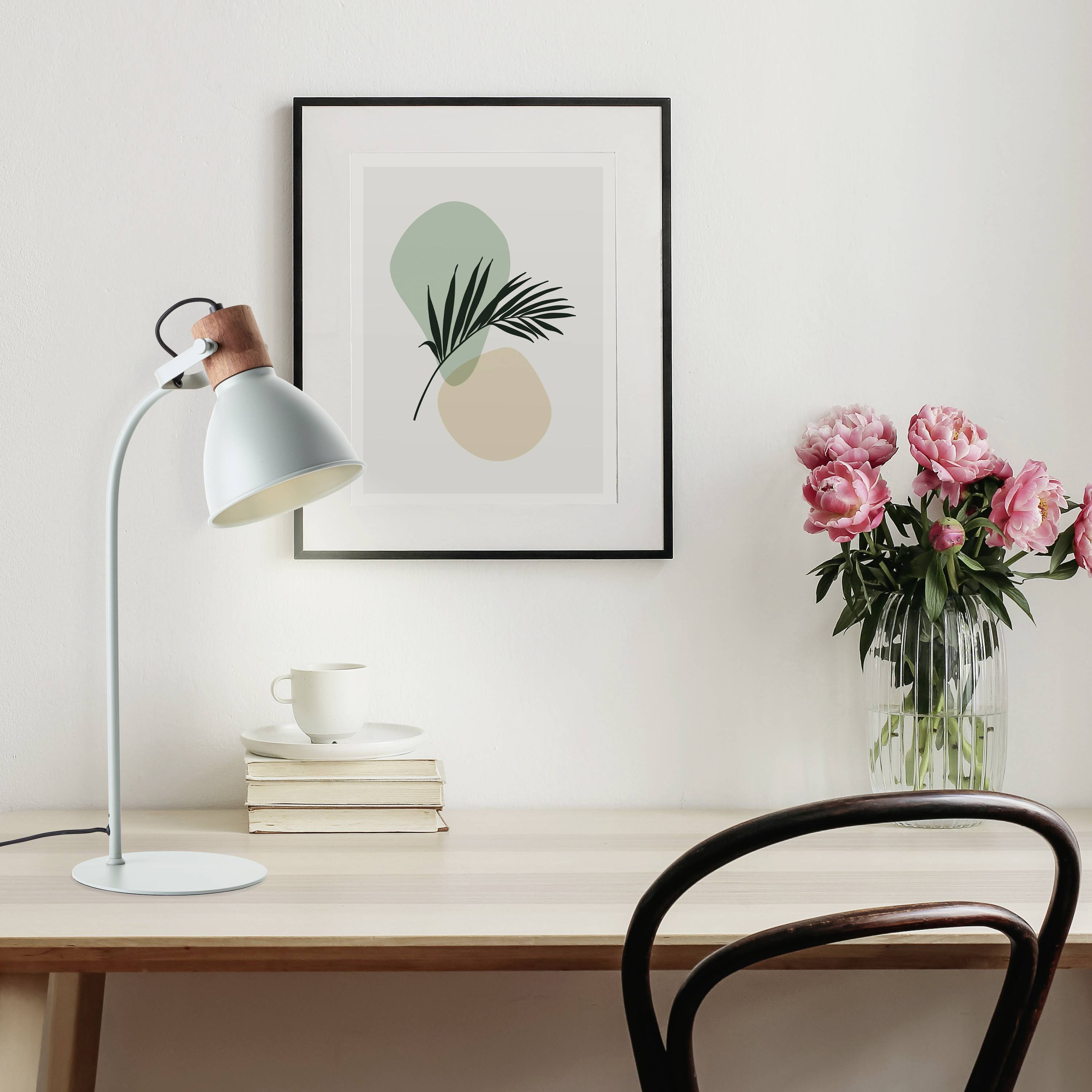 Ein Schreibtisch mit einer Lampe, einem Buchstapel und einer Tasse. An der Wand hängt ein minimalistisches Bild. Rechts steht eine Vase mit rosa Blumen.