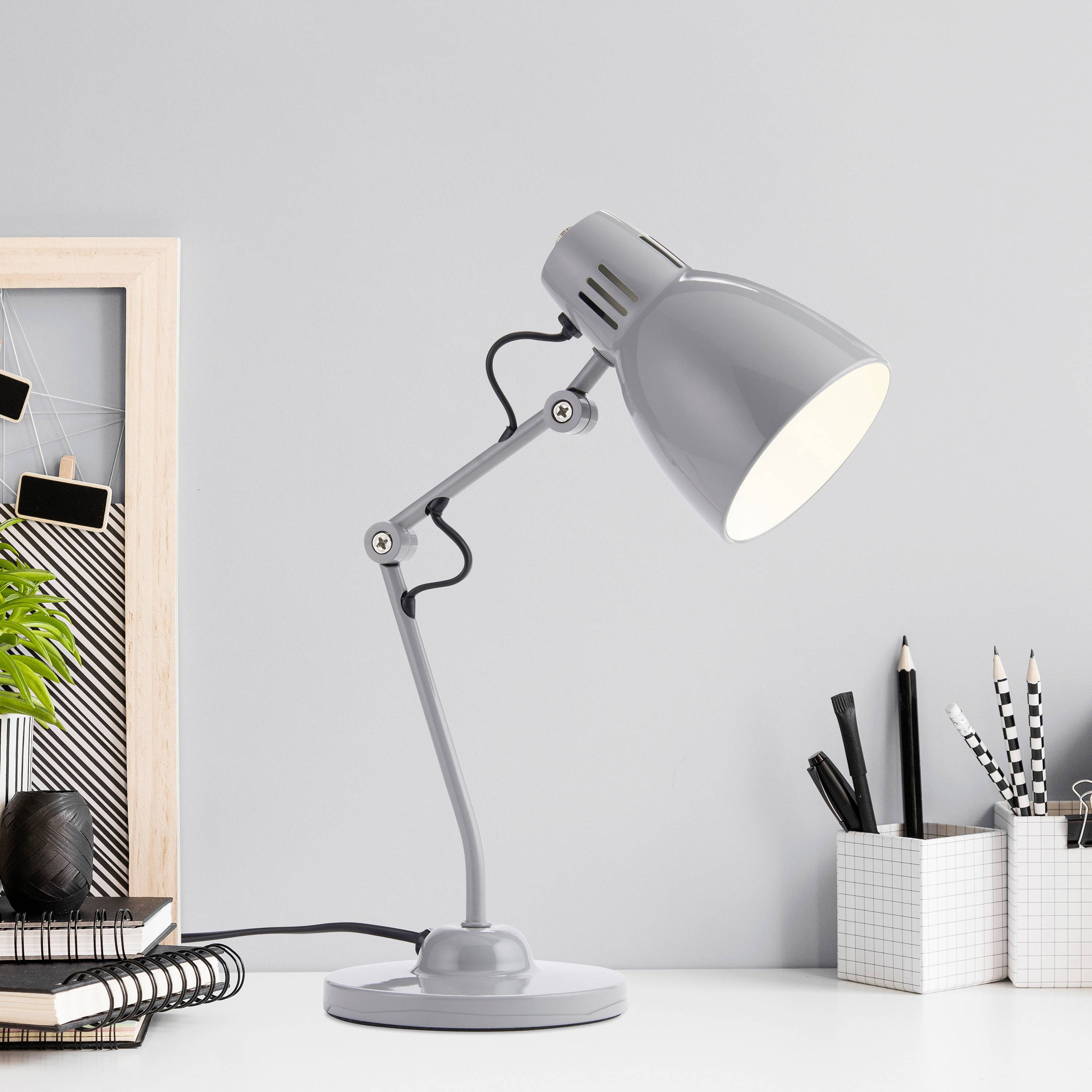 Tischlampe auf Schreibtisch, strahlt Licht auf Notizbuch und Stiftehalter. Rahmen mit Pflanze im Hintergrund.