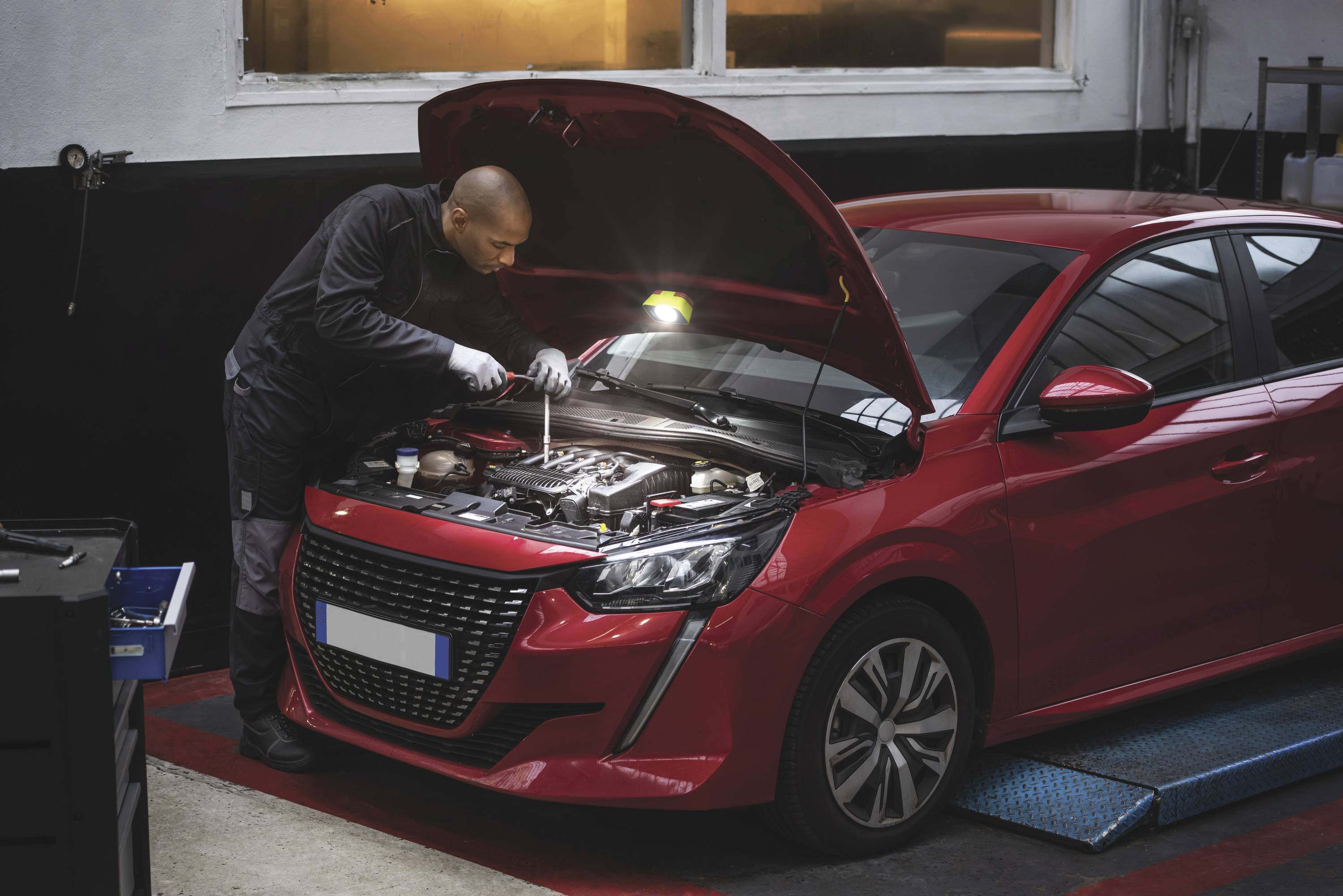 Ein Mechaniker repariert einen roten Kleinwagen mit geöffneter Motorhaube in einer Autowerkstatt. Fokus auf den Motor.