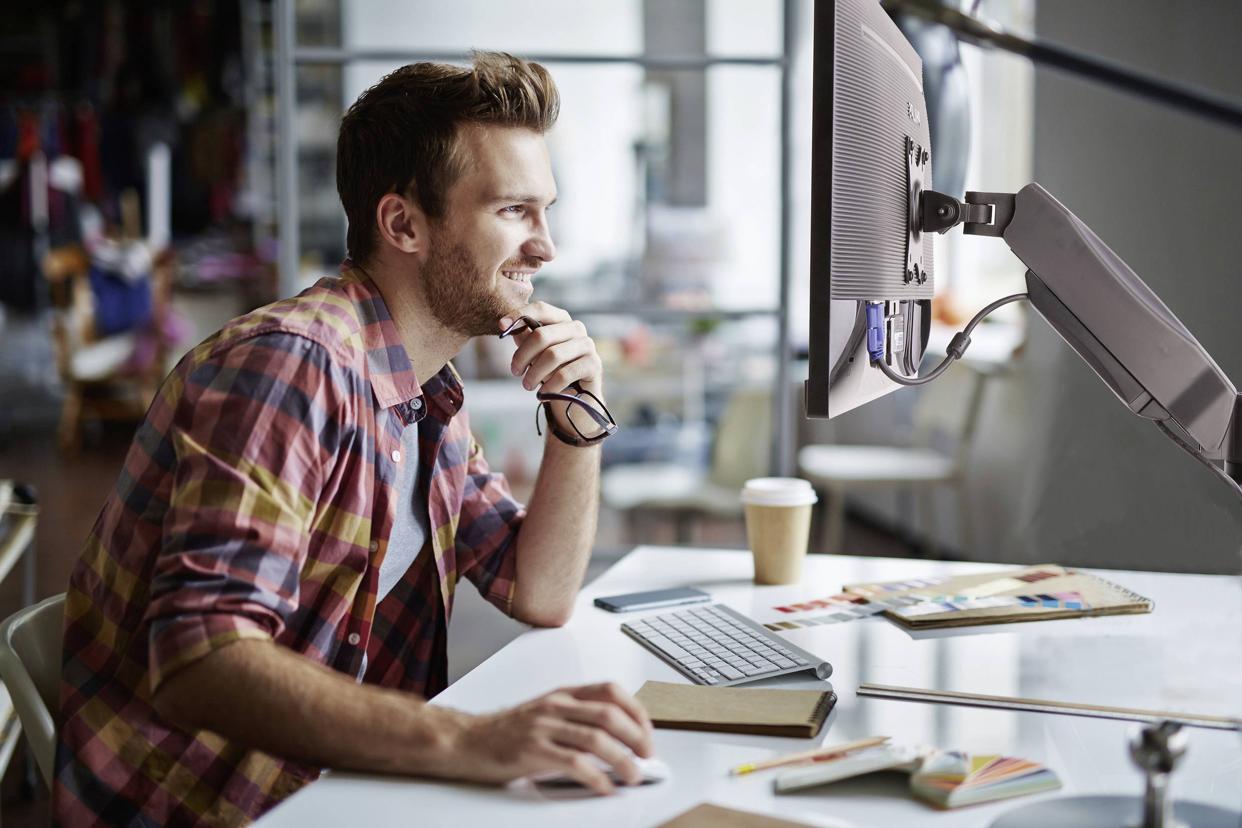 Ein Mann sitzt lächelnd an einem Schreibtisch vor einem Computerbildschirm, umgeben von Notizbüchern und Zeichenmaterial.
