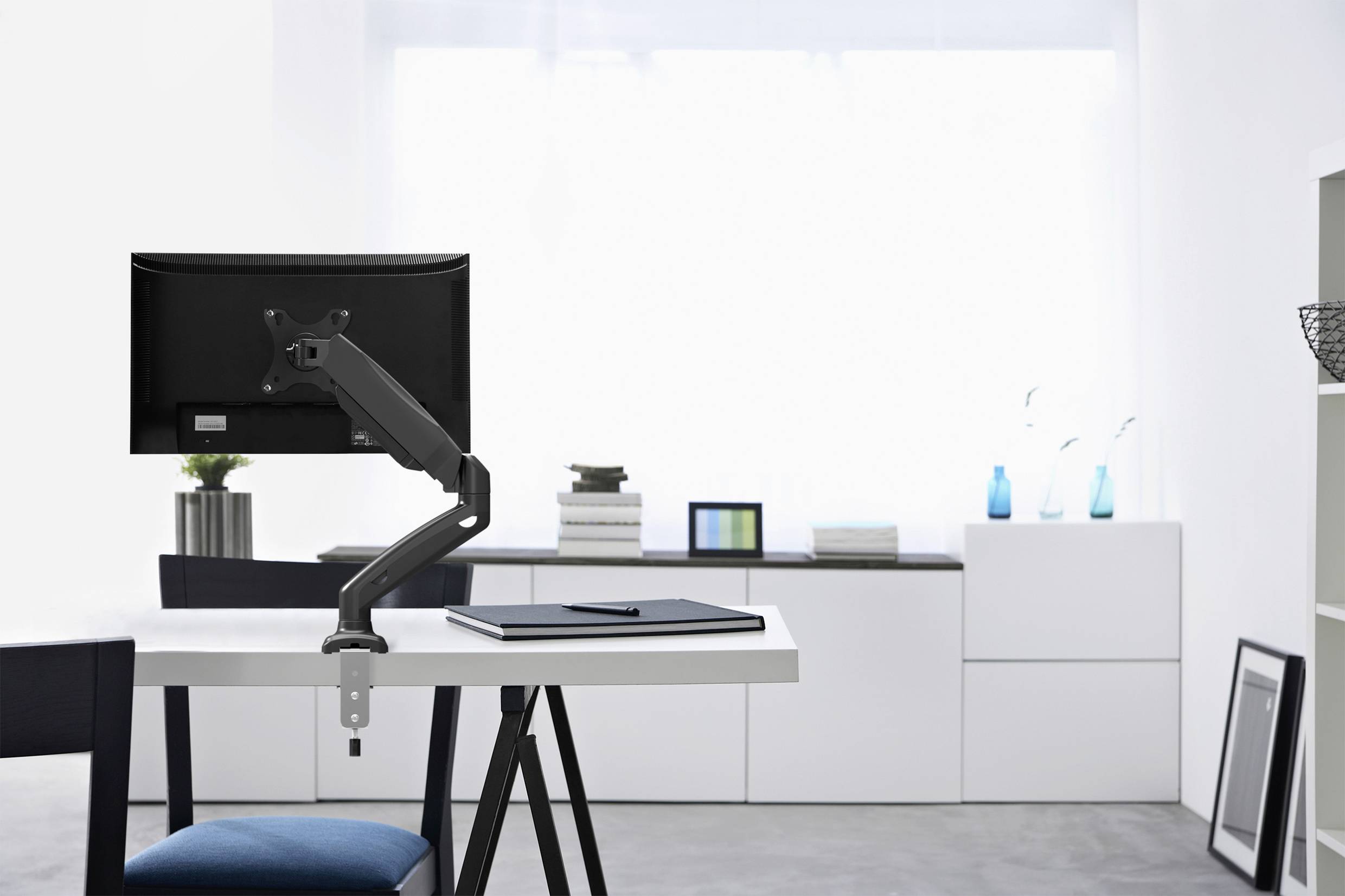 Moderner Arbeitsplatz mit einem schwarzen Monitor an einem Schwenkarm. Weiße Möbel und Dekor im Hintergrund. Helle, minimalistische Einrichtung.