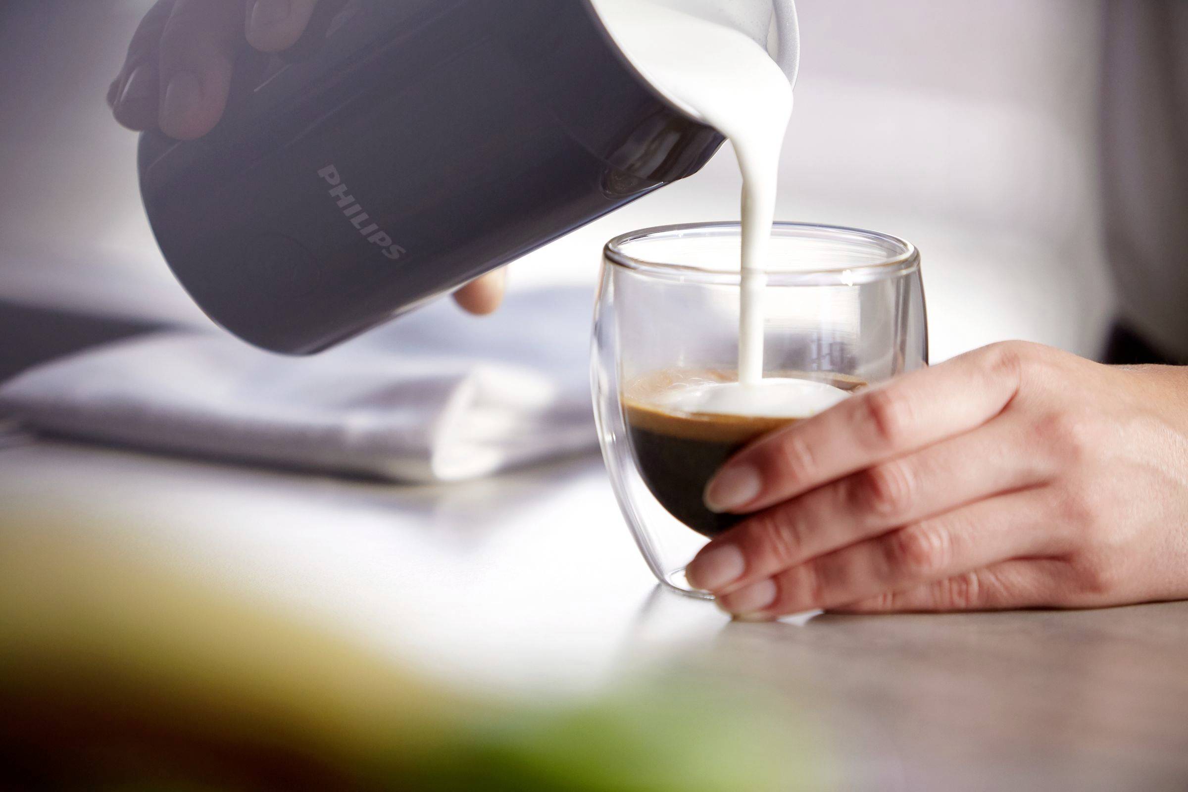 Eine Person gießt Milch aus einem Krug in ein Glas mit Kaffee auf einem Tisch.
