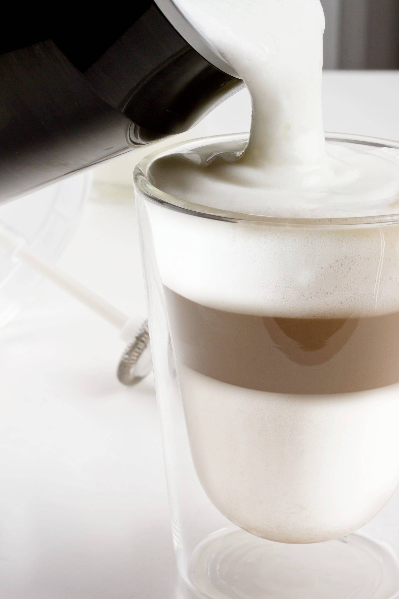 Ein Glas Latte Macchiato wird mit aufgeschäumter Milch aus einem Krug gefüllt. Drei Schichten sichtbar: Milch, Kaffee, Schaum.