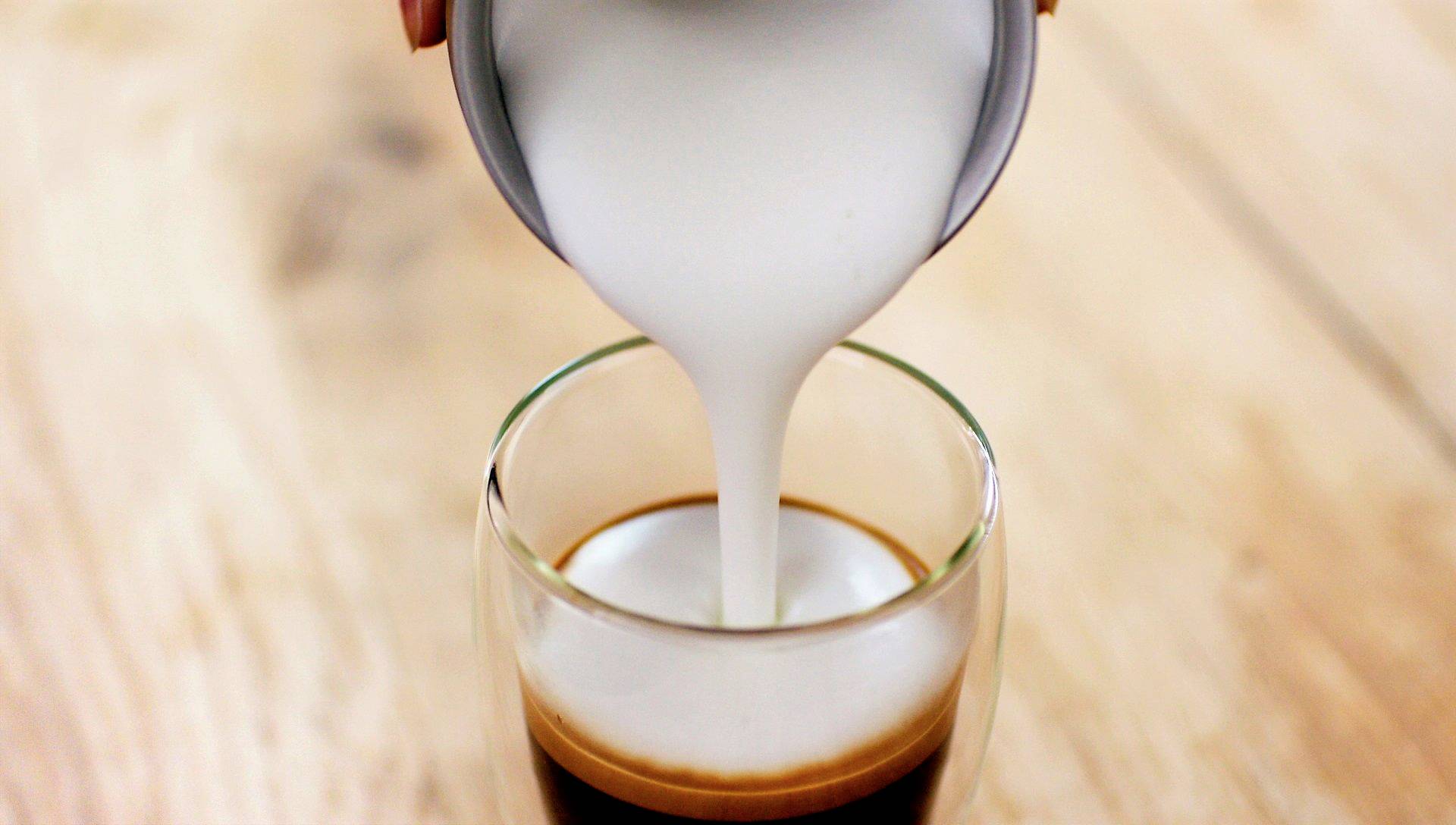 Eine Person gießt aufgeschäumte Milch in ein Glas mit Espresso, um einen Latte Macchiato zuzubereiten, auf einem Holztisch.