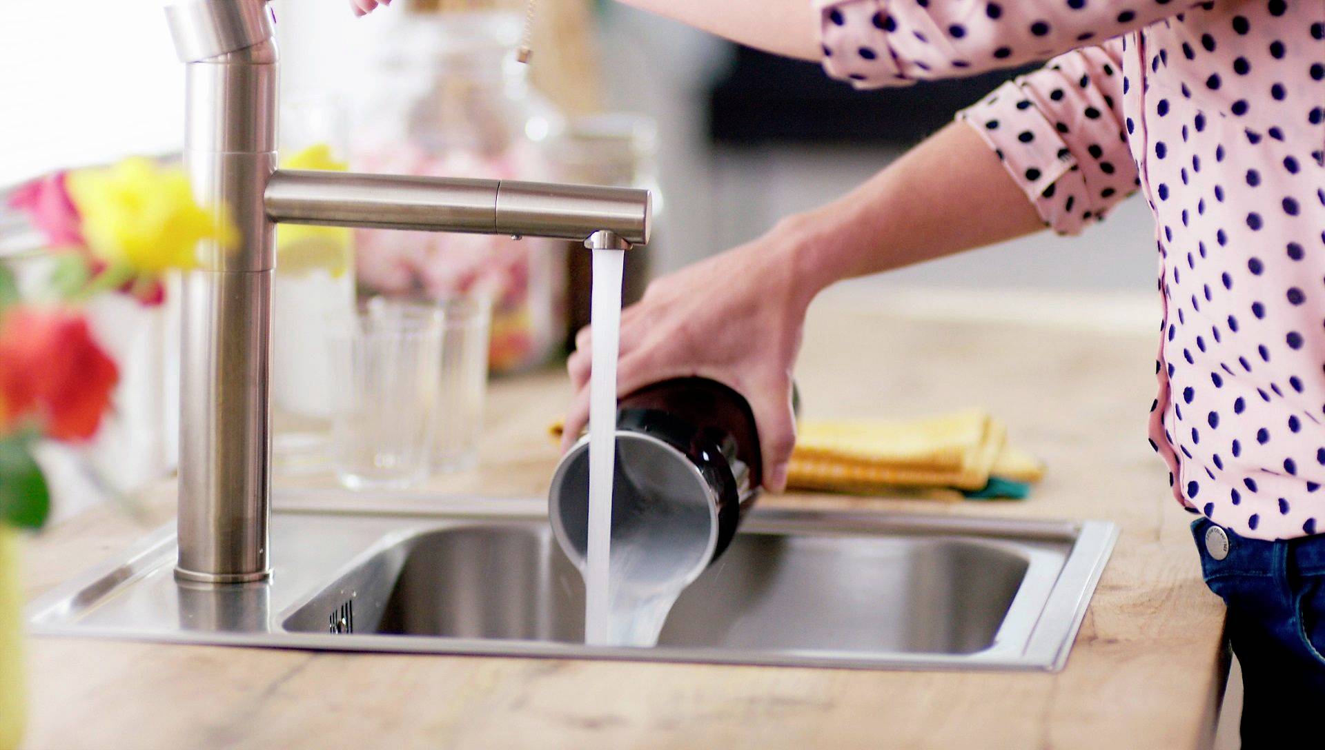 Eine Person spült ein Glas unter fließendem Wasser in einem Küchenspülbecken. Im Hintergrund sind Blumen und ein gelbes Geschirrtuch.