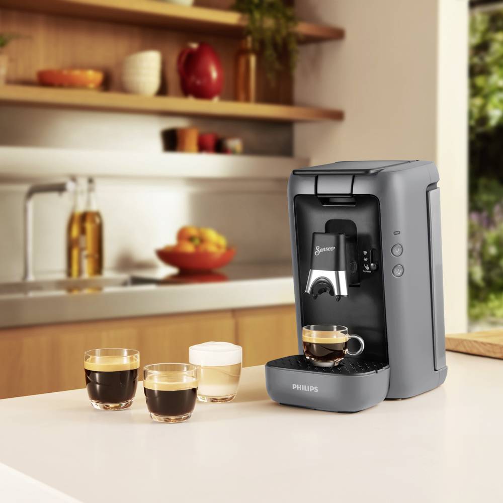 Eine Kaffeemaschine auf einer Küchenarbeitsplatte bereitet Espresso. Daneben stehen zwei Tassen Espresso und eine Tasse Milchschaum.