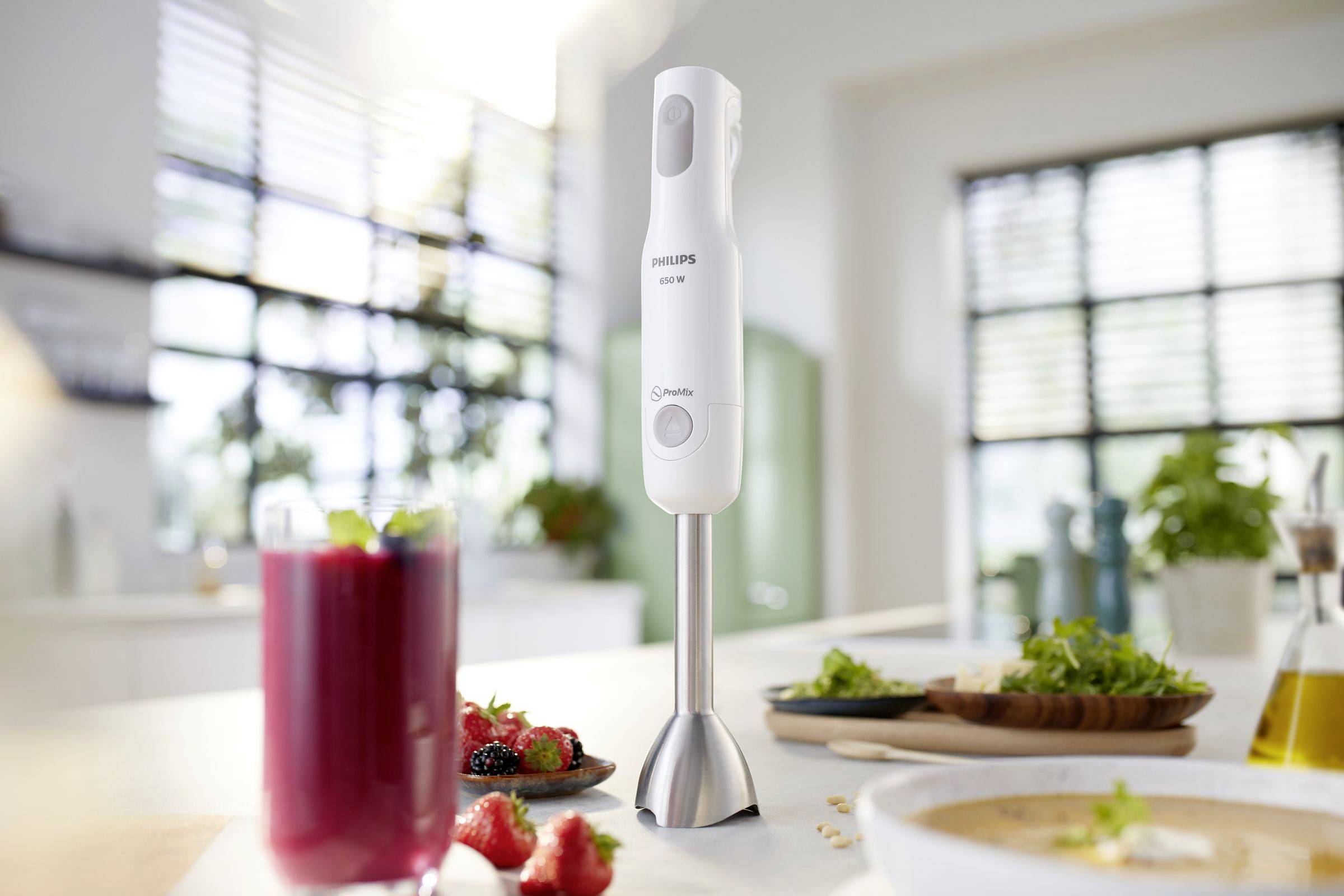 Ein weißer Stabmixer steht auf einer Küchentheke. Im Vordergrund ein Glas mit rotem Smoothie und frischem Obst und Gemüse.