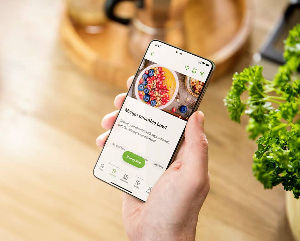 Eine Hand hält ein Smartphone mit einer App, die ein Rezept für einen 'Mango Smoothie Bowl' zeigt. Im Hintergrund ist Petersilie zu sehen.