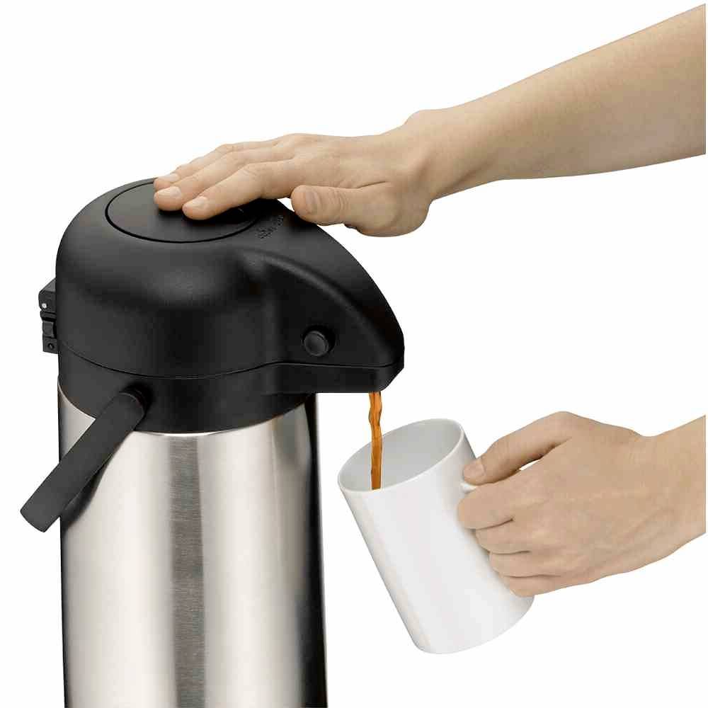 Eine Hand drückt auf den Hebel eines silbernen Thermoskanne-Spenders, während eine andere Hand eine weiße Tasse darunter hält, in die Flüssigkeit fließt.