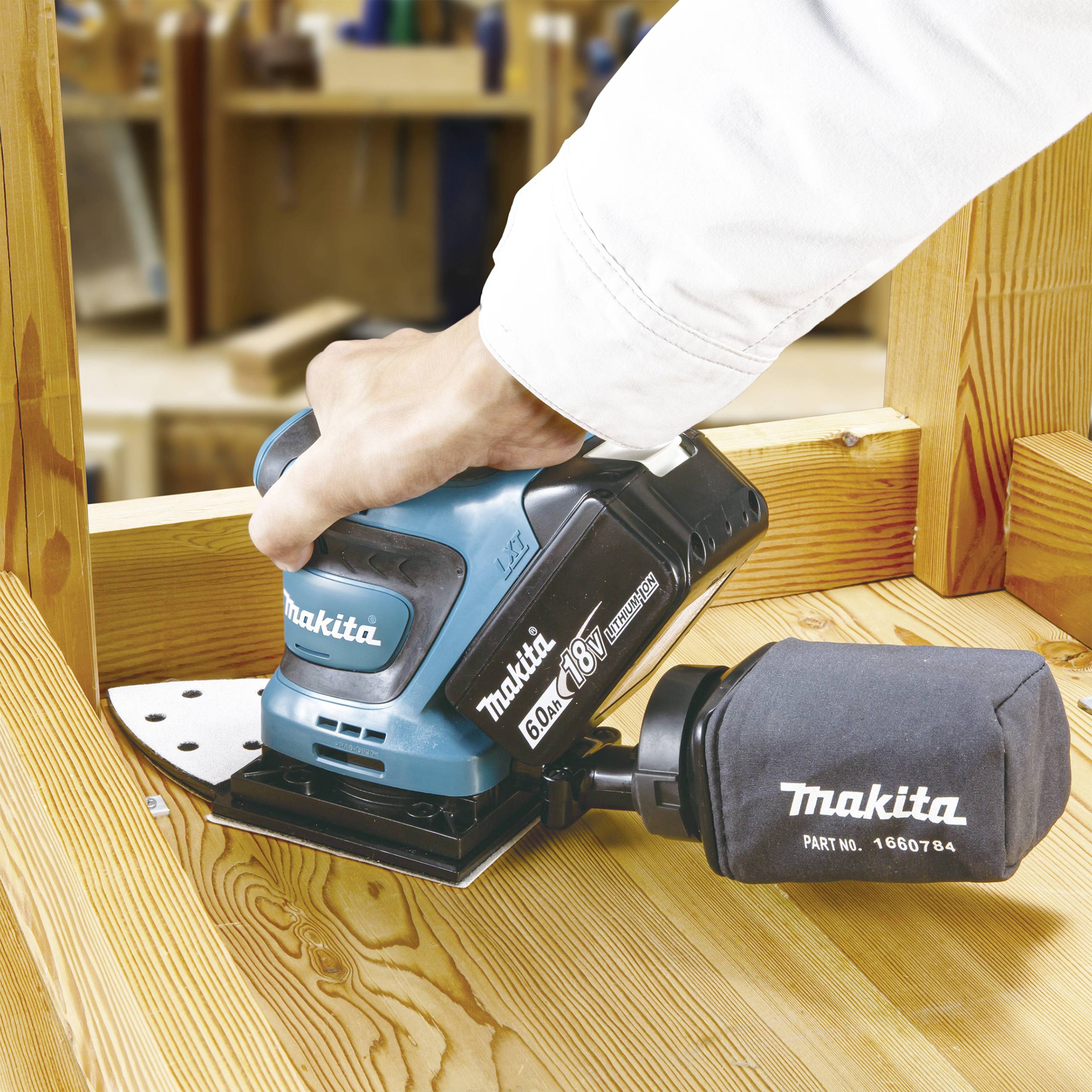 Ein Handwerker nutzt einen blauen Makita-Schleifer, um ein Holzbrett zu schleifen. Im Hintergrund sind Werkzeuge sichtbar.