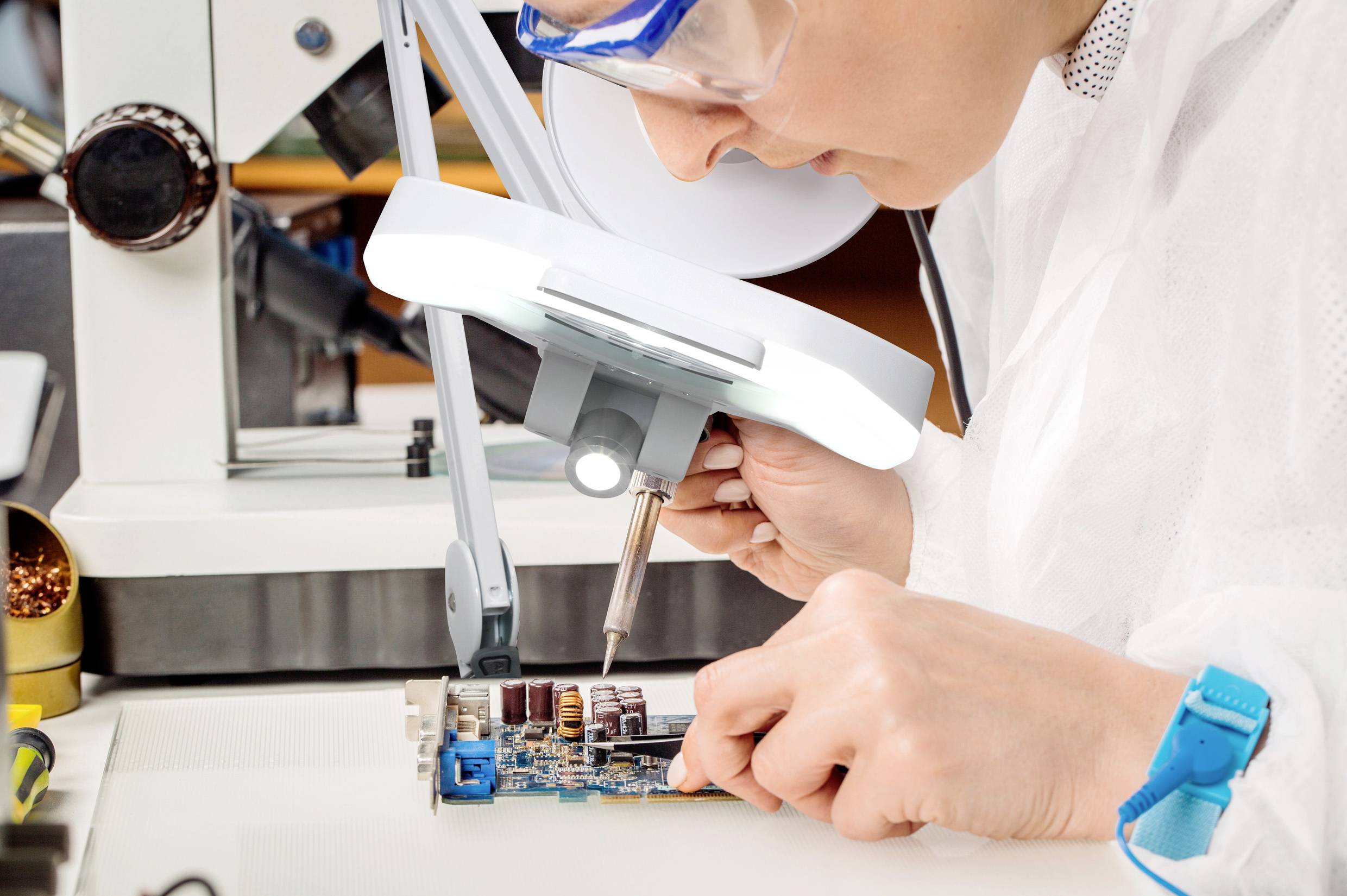 Eine Person in einem Labor arbeitet unter einer Lupe an einer Leiterplatte und verwendet dabei ein Lötkolben, um Bauteile zu befestigen.