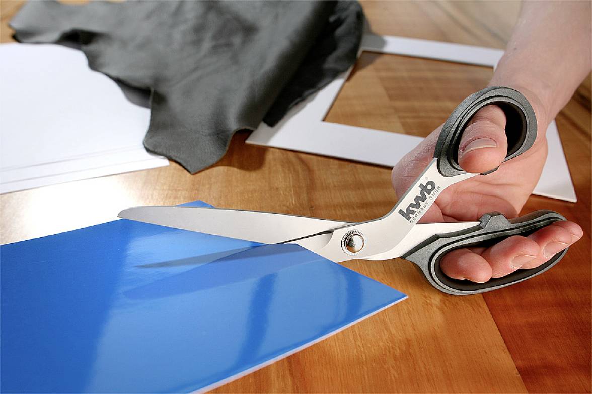 Eine Hand verwendet eine große Schere, um ein blaues Blatt Papier auf einem Holztisch zu schneiden. Im Hintergrund liegen Stoff und Papier.