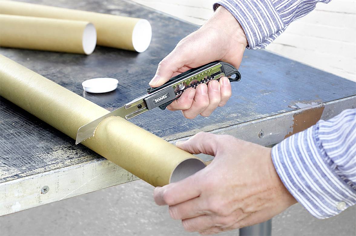 Eine Person schneidet mit einem Cuttermesser ein braunes Papprohr auf einem Metalltisch. Hände und Messer sind sichtbar.
