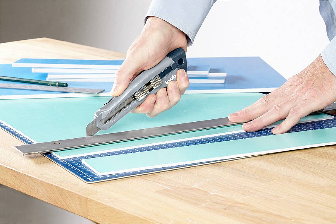 Jemand schneidet mit einem Cutter und Lineal durch mehrere blaue und grüne Kunststoffplatten auf einem Holztisch.