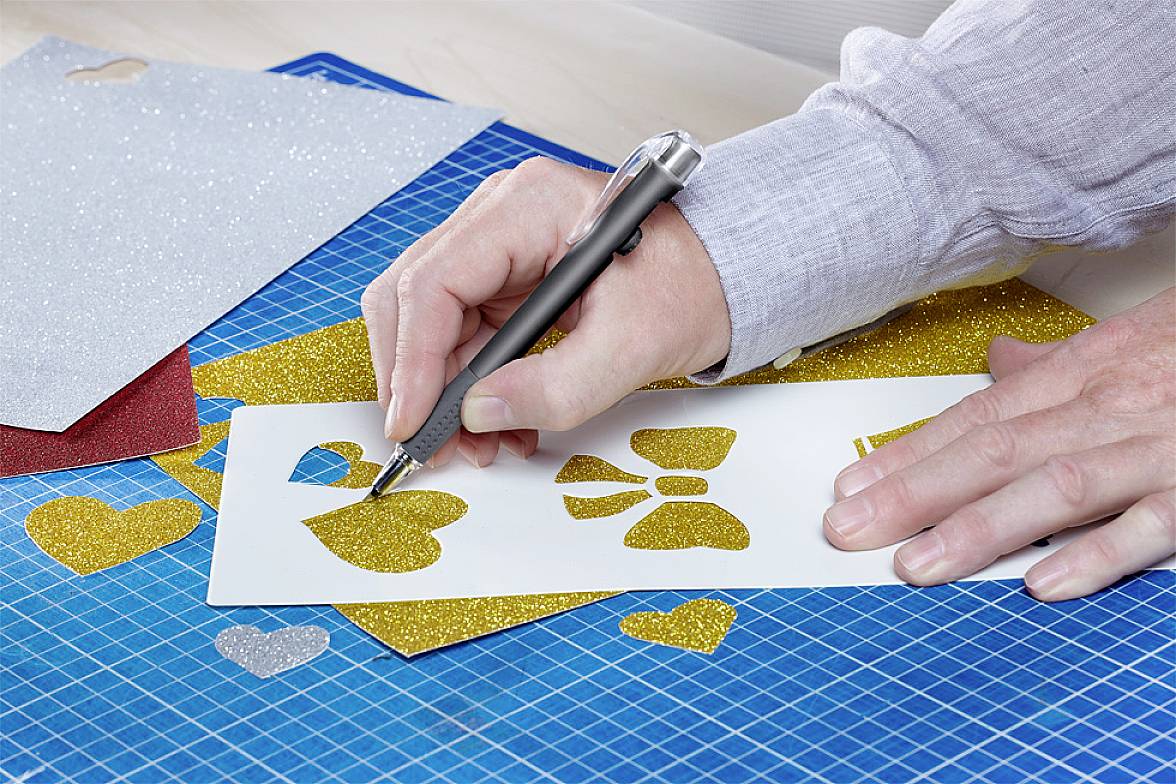 Person schneidet goldene Formen aus Glitzerpapier mit einem Skalpell auf einer Schneidematte aus. Formen: Herzen, Schleifen.