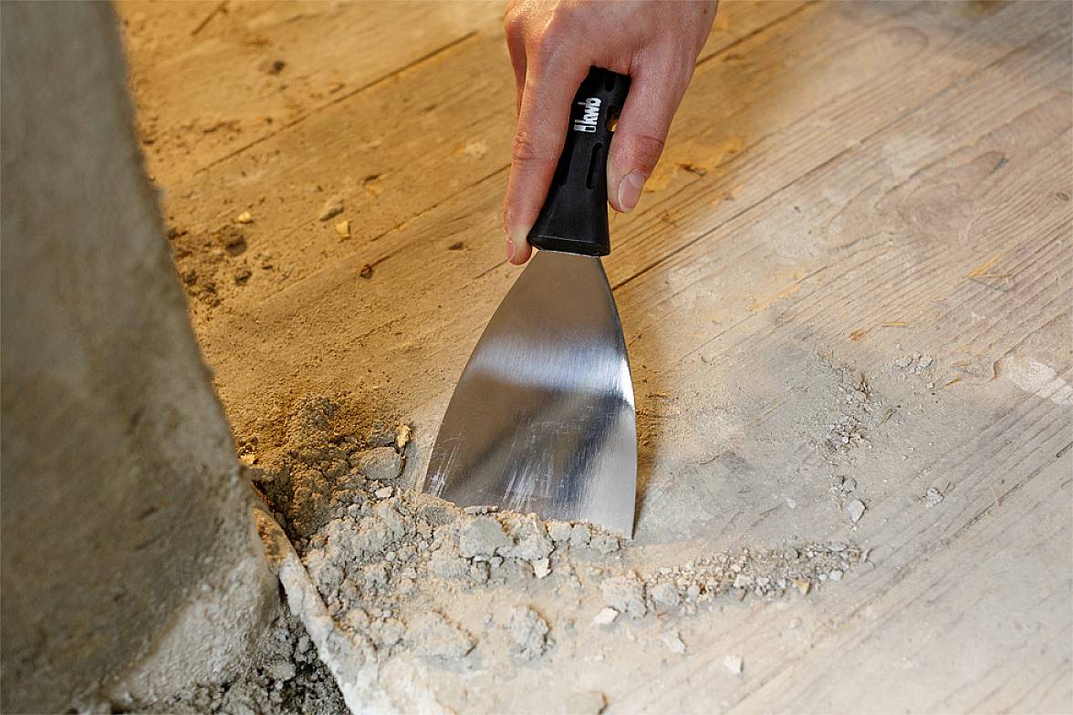 Eine Hand hält einen Spachtel und entfernt alten Mörtel von einem Holzboden an der Kante eines Betonsockels.