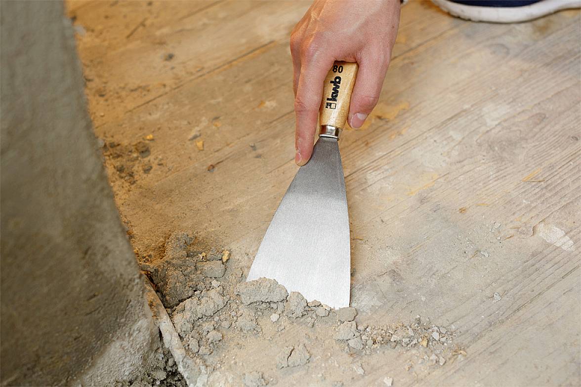 Eine Hand benutzt einen Spachtel, um trockenen Mörtel von einem Holzboden in der Nähe einer Wand zu entfernen.