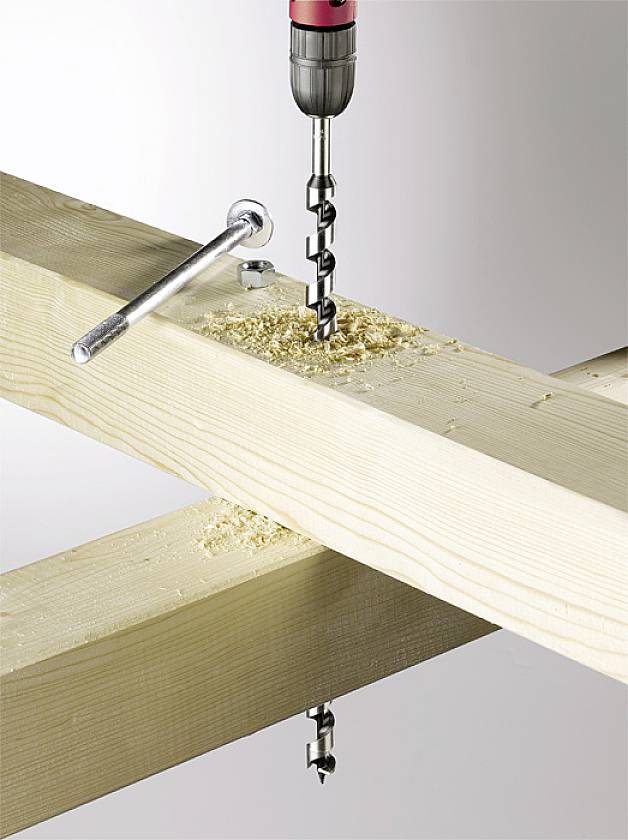 Eine Bohrmaschine bohrt ein Loch in einen Holzbalken. Holzspäne sind um das Loch verteilt. Ein Schraubenschlüssel liegt daneben.